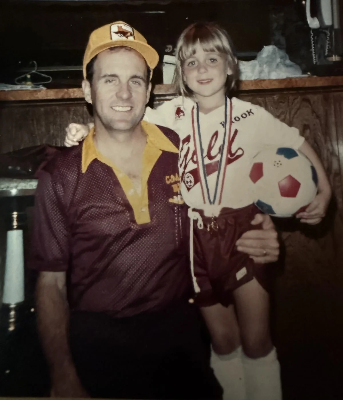 A man and a young girl posing together indoors. The girl is holding a soccer ball and wearing a medals around her neck. The man is wearing a yellow cap and a maroon shirt, both with sports team logos. The girl is dressed in a sports uniform. They are smiling at the camera.