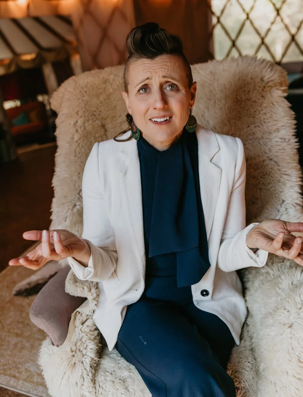 A woman sitting in a plush, cream-colored chair with a distressed, confused expression, gesturing with her hands.
