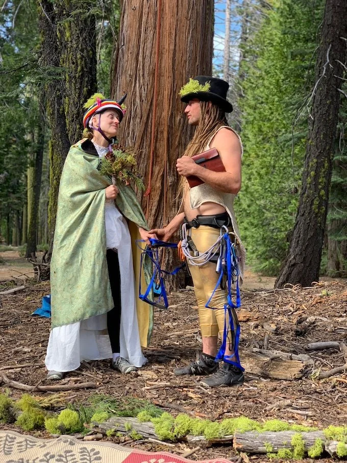Two people dressed in fantasy costumes stand in a forest, holding hands in front of a large tree. One is dressed as a fairy or elf with a colorful hat, green cape, and holding a bouquet of flowers. The other is dressed as a witch or wizard with a black hat, holding a book, and wearing yellow pants with climbing gear attached.