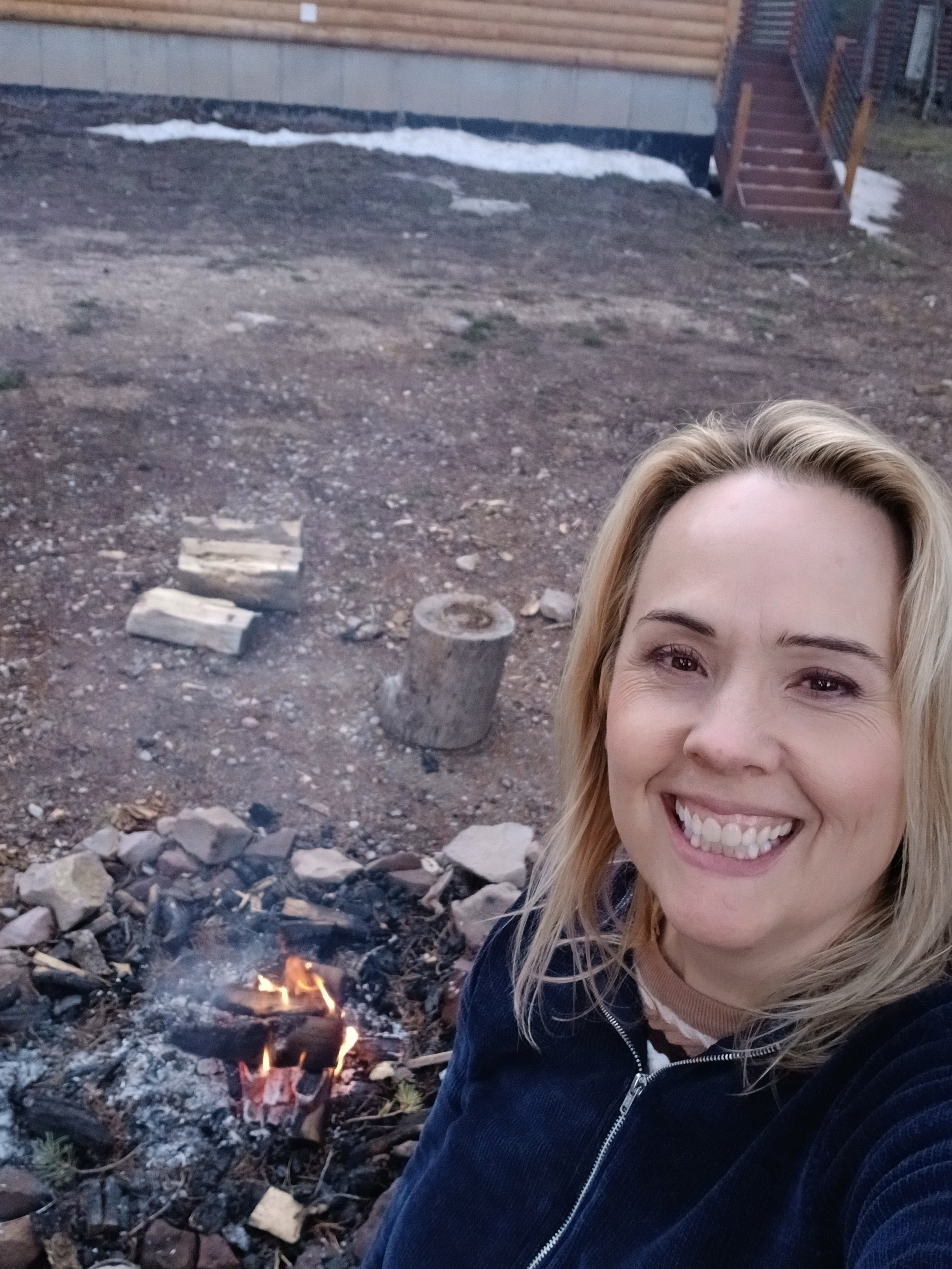 A smiling woman with blonde hair taking a selfie outdoors near a small campfire, with a wooden house and stairs in the background.