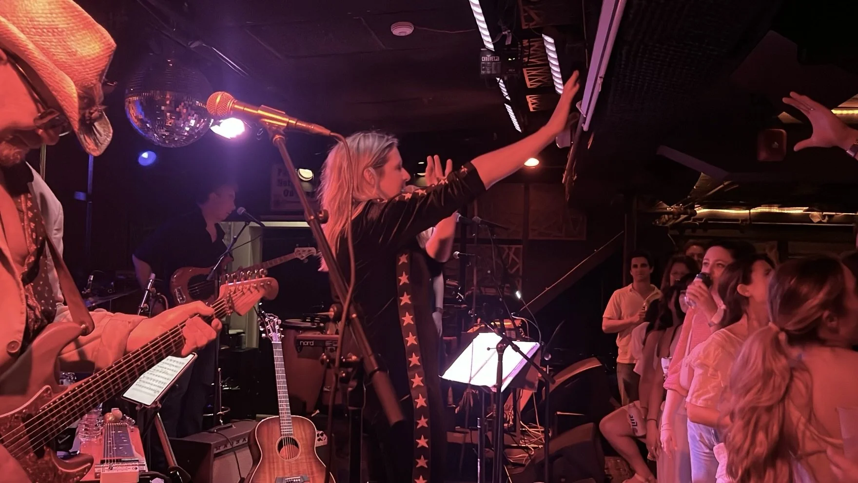 A live music performance at a small venue with a band playing on stage and a woman in front of the stage reaching up towards the equipment, with audience members watching and some drinking.
