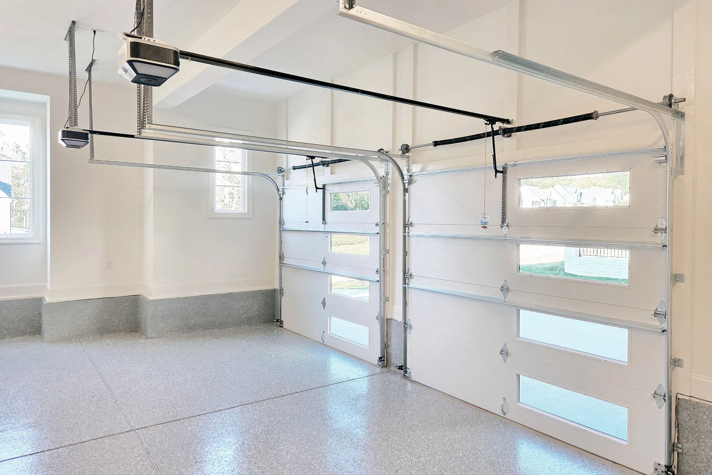 Empty garage with white garage doors featuring small windows and automatic openers, and a speckled epoxy floor.