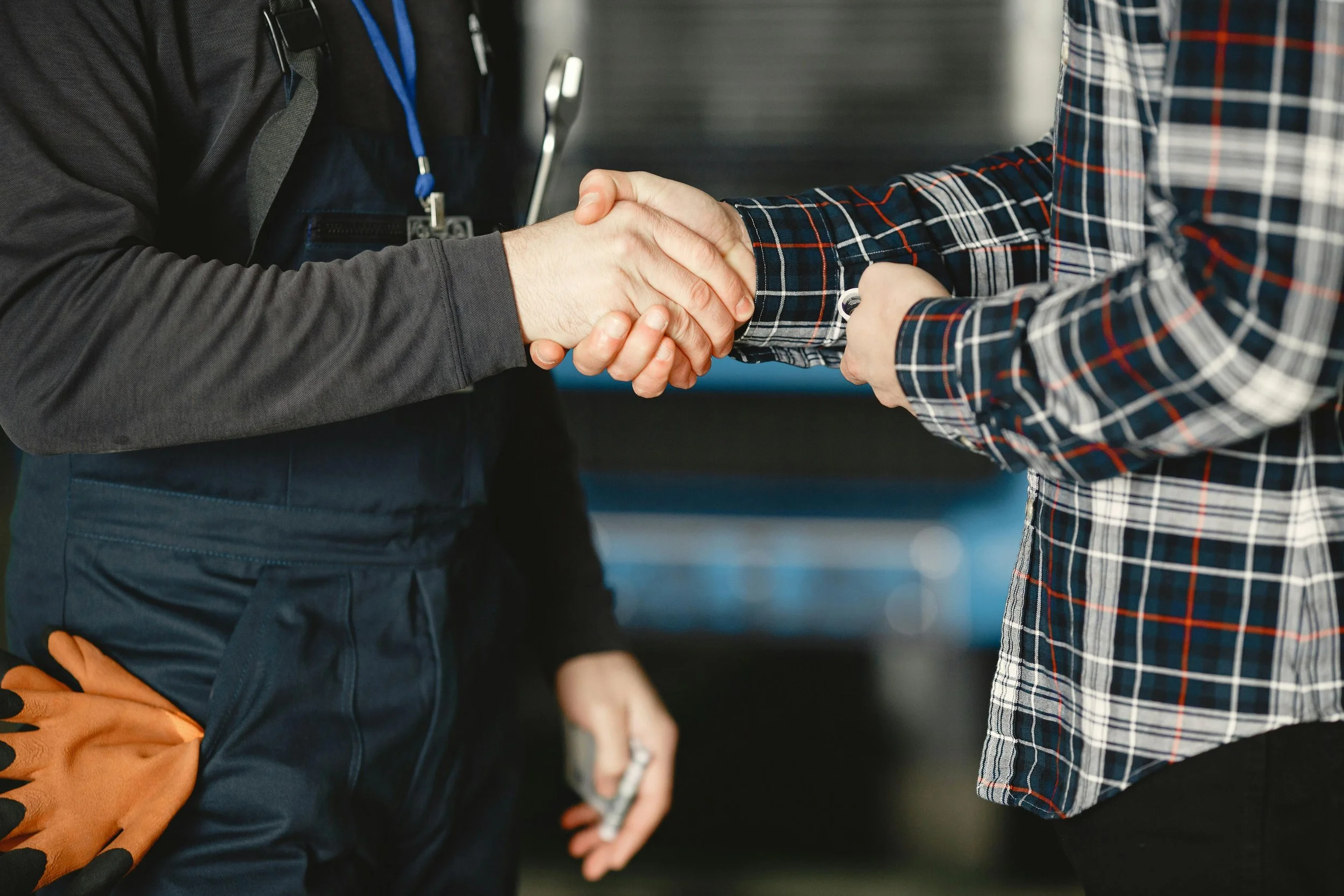 Two people shaking hands in an indoor setting, one wearing a dark uniform with a tool hanging from the pocket and the other in a checkered shirt, possibly indicating a professional or business interaction.