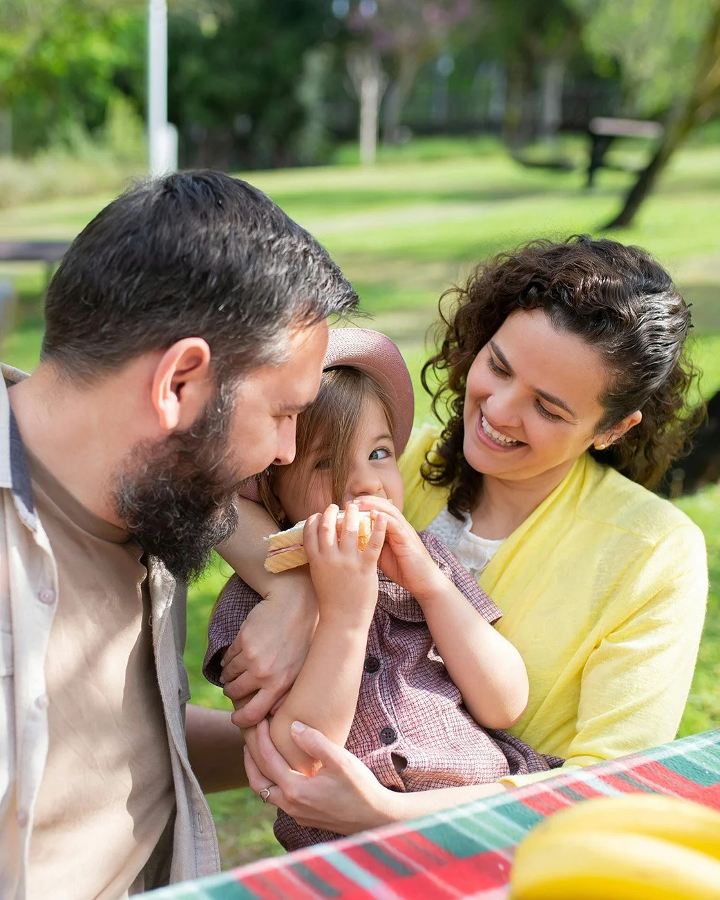 A family of three, a man, a woman, and a young girl, sharing a moment outdoors at a park during a picnic. The girl is eating a sandwich and smiling while being held by the woman, with the man leaning in.