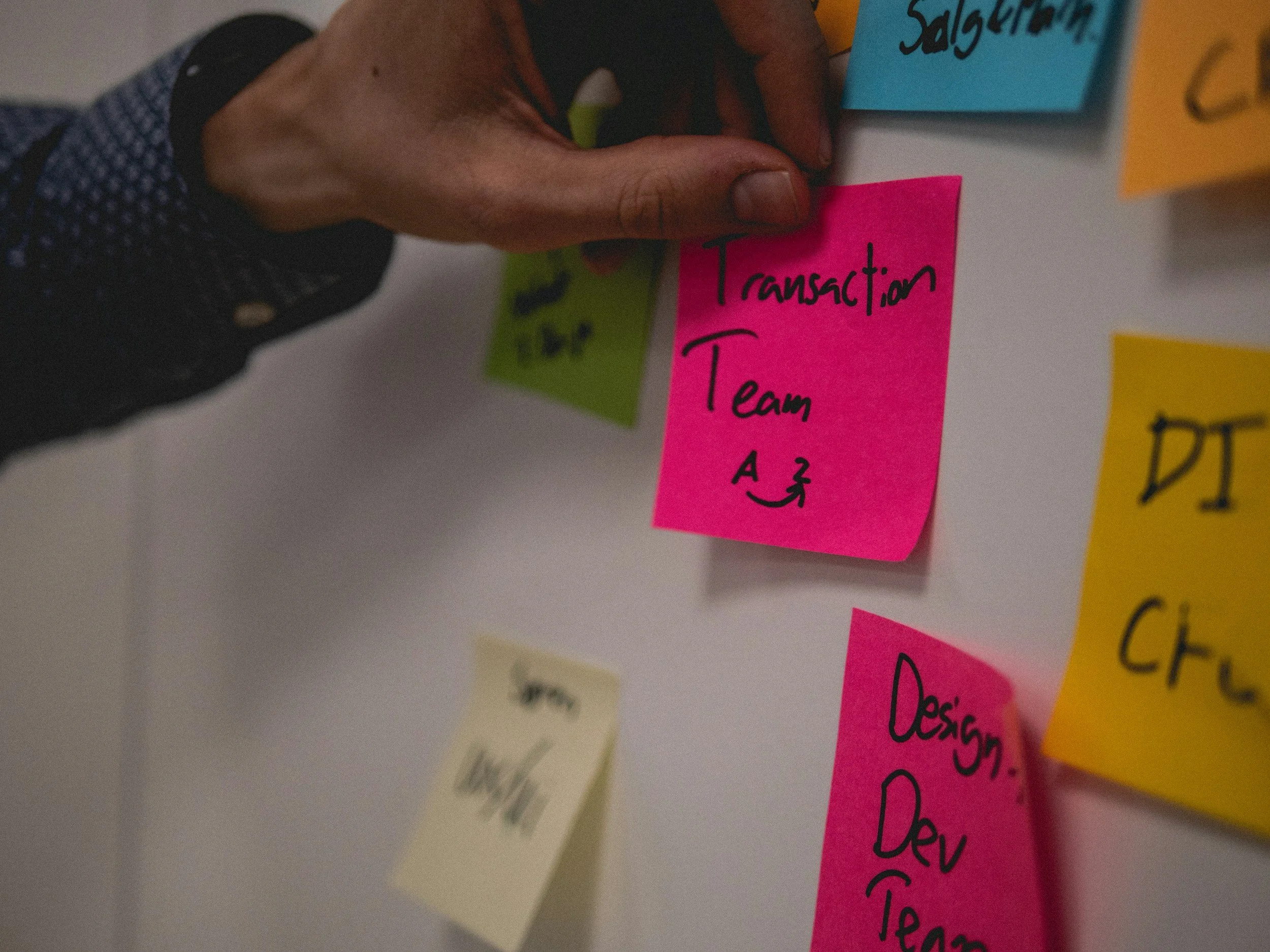 A person's hand placing or adjusting a bright pink sticky note on a wall covered with other colorful sticky notes. The pink sticky note has handwritten text: "Transaction Team" with an arrow below. Other notes include a yellow note that says "DI" and a blue note near the top right. The person is wearing a dark, patterned long sleeve shirt.
