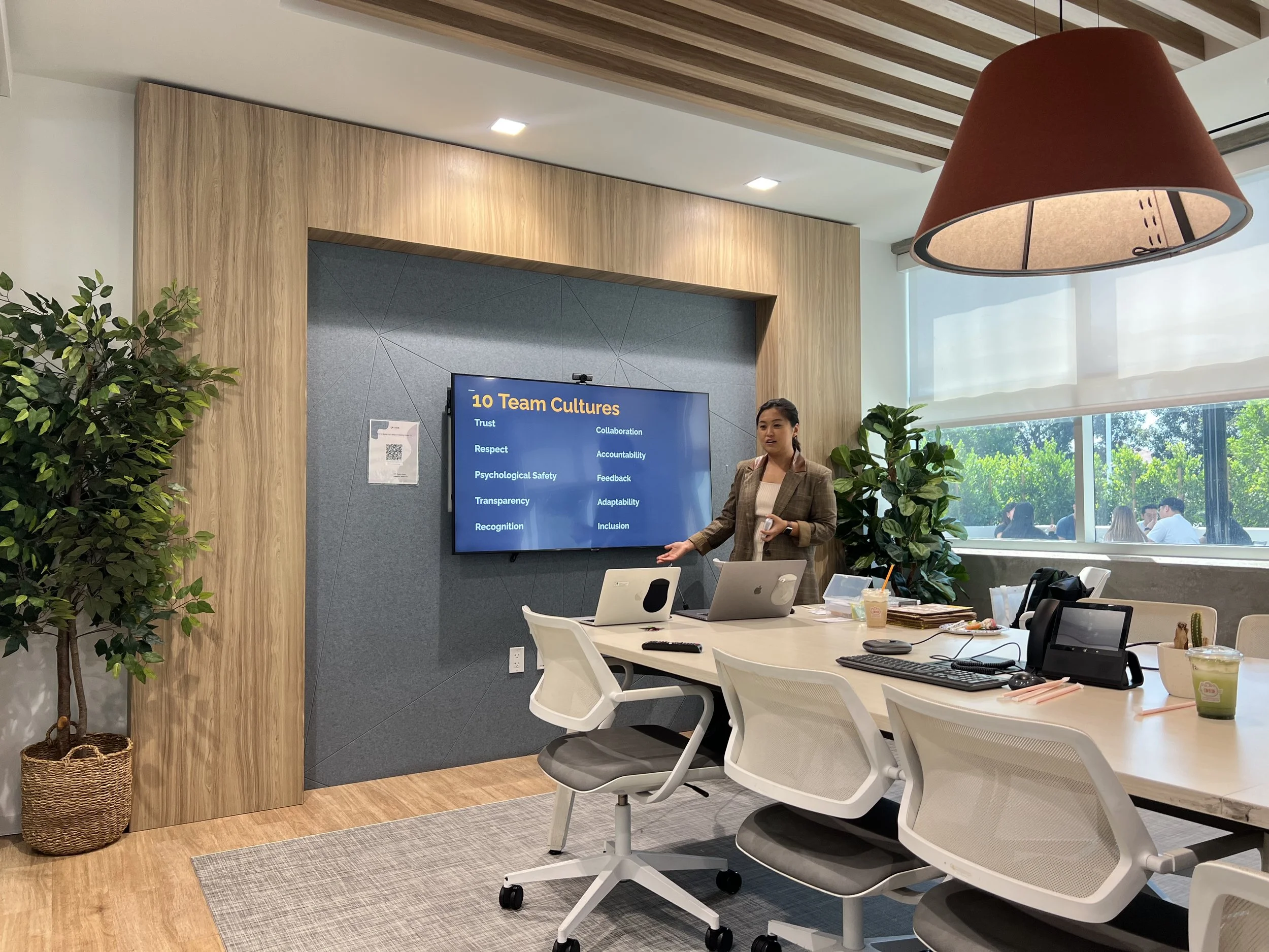 Woman giving a presentation on team cultures in a modern office conference room with a large window, plants, and electronic devices on the table.