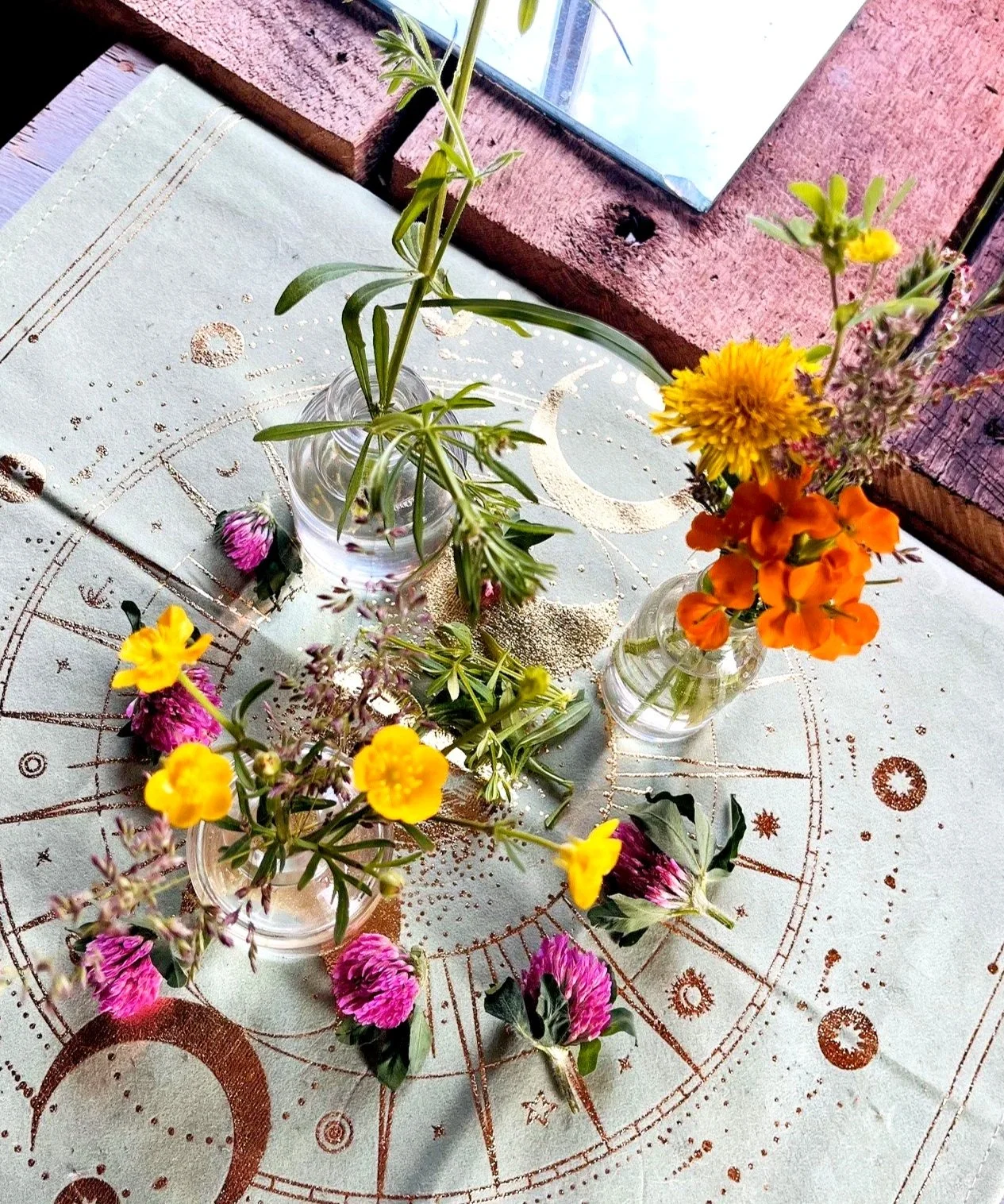 Four small bottles with colorful flowers, including yellow, orange, pink, and purple, placed on a cloth with celestial and star patterns. The scene is set near a window with a wooden frame.