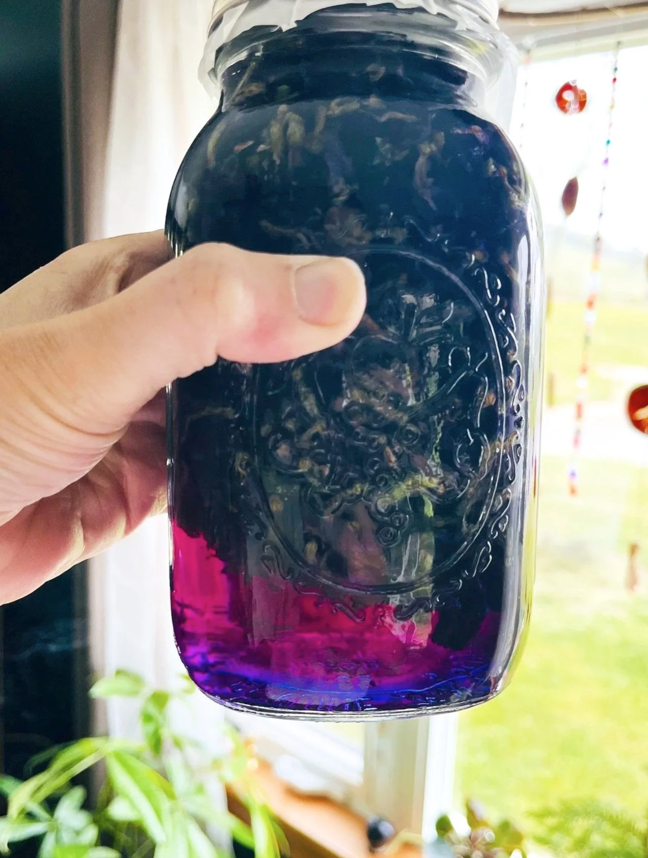 A hand holding a glass jar with a colorful layered liquid, transitioning from purple at the bottom to pink and dark blue at the top.