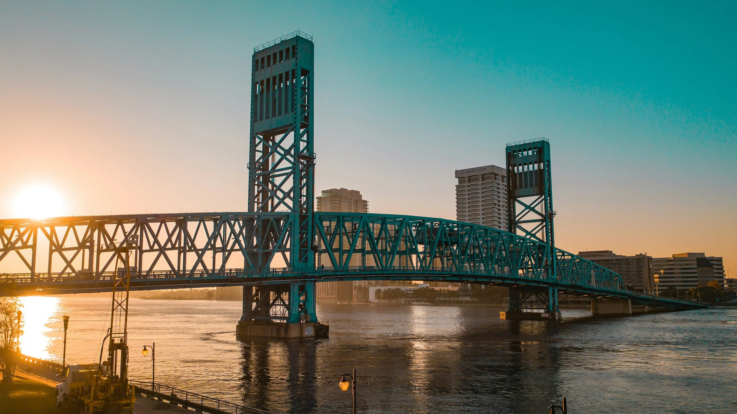 Jacksonville  cityscape at sunset with a blue steel bridge over a river and tall buildings in the background.