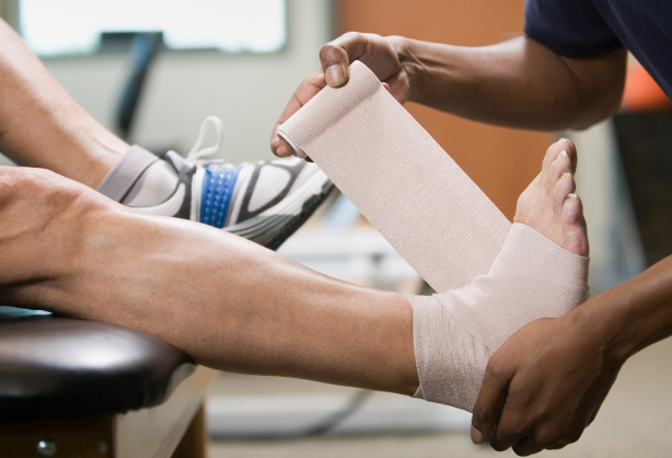 A person in a black shirt applies a beige bandage to another person's lower leg, which is resting on a table, in a medical or physiotherapy setting.