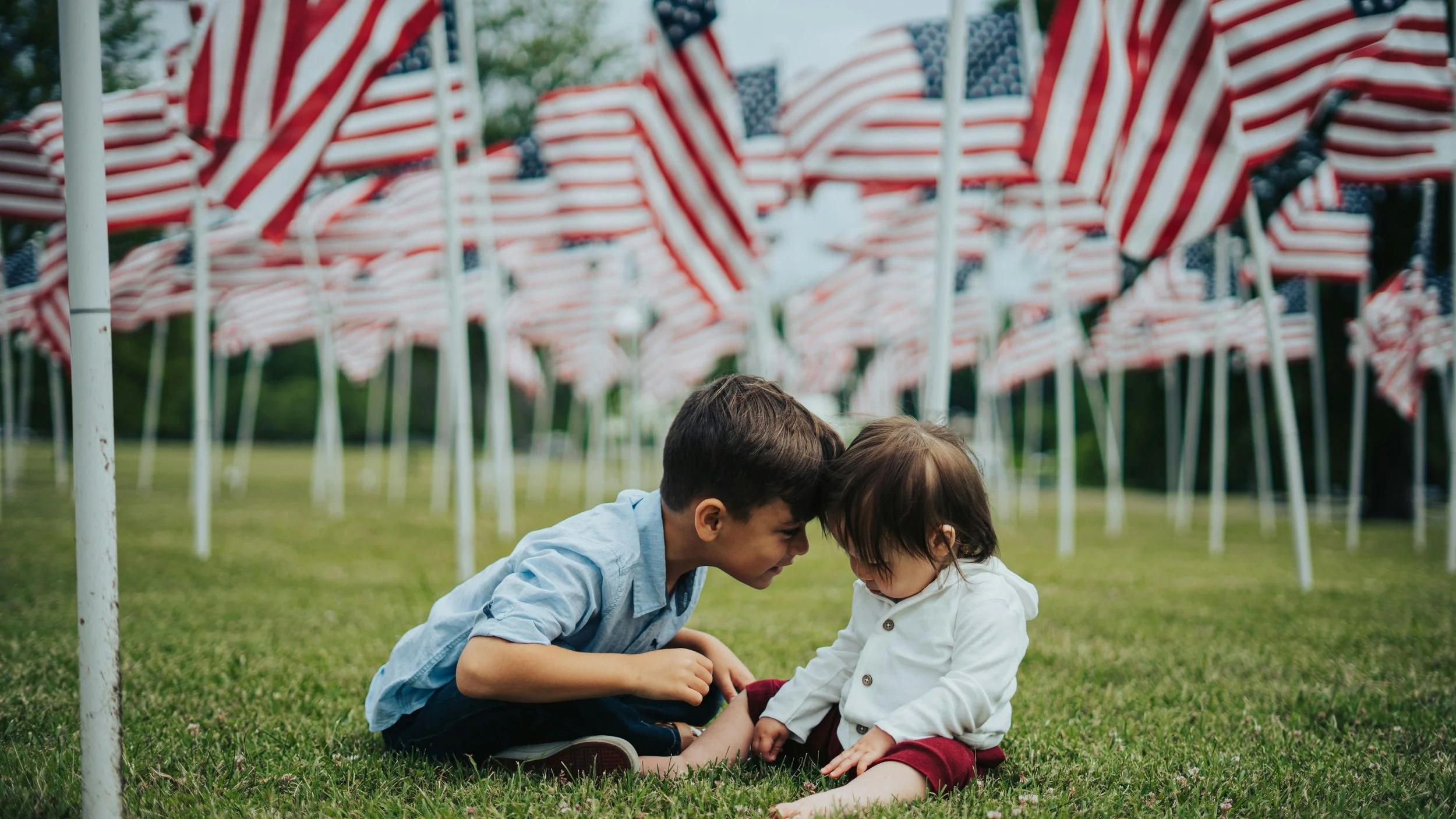 A young man and a young girl are sitting on the grass under rows of American flags, touching foreheads and looking at each other.