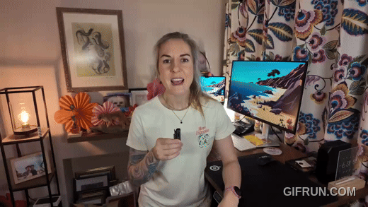 A young YouTube woman with long blonde hair and tattoos on her arms standing in a decorated room, holding a small mic and speaking, with two monitors, a desk, and colorful curtains in the background.