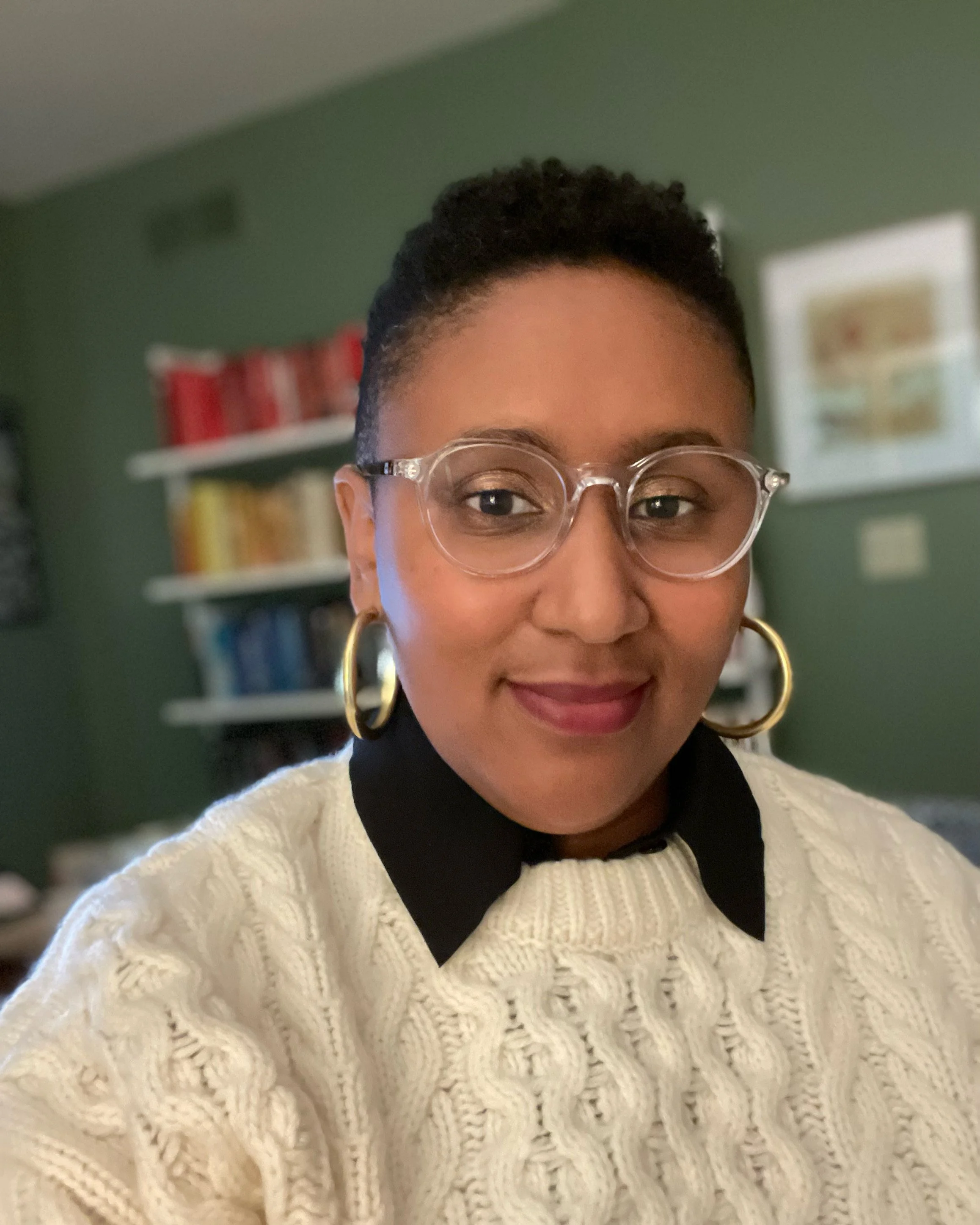A woman with short black hair, wearing clear glasses with round frames, large gold hoop earrings, a black shirt, and a cream-colored textured sweater, smiling indoors with a green wall and a bookshelf in the background.
