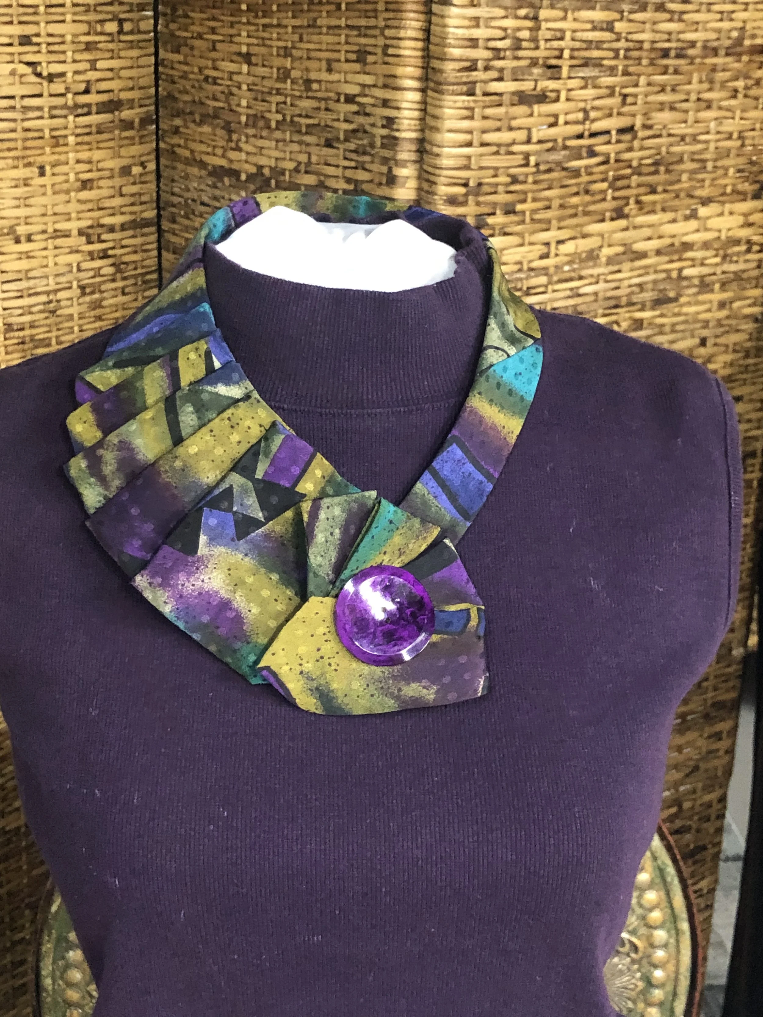 A mannequin dressed in a purple top wearing a colorful, abstract-patterned scarf with a large purple circular brooch, set against a woven wicker background.