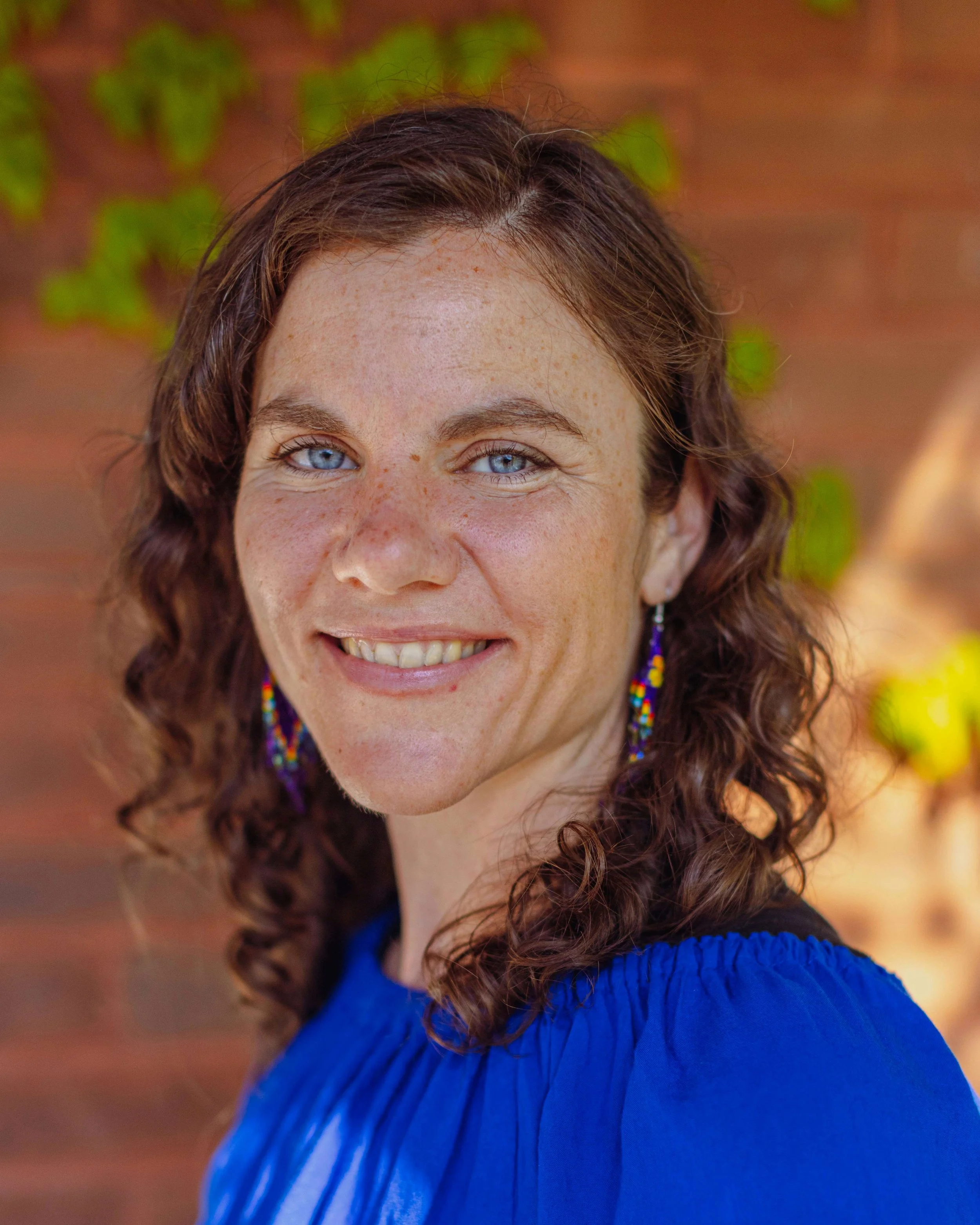 Close-up of a smiling woman with curly brown hair, blue eyes, freckles, wearing earrings and a blue sweater, outdoors with a blurred background of green and brown foliage.