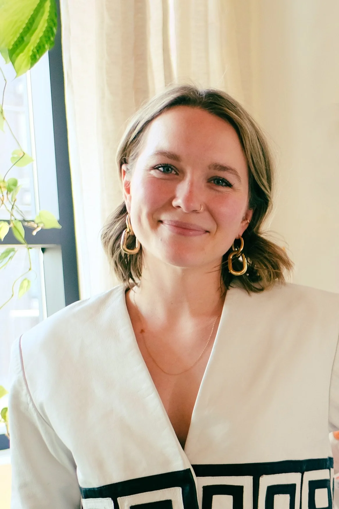 A young woman with short brown hair, light skin, and blue eyes is smiling in front of a window with beige curtains and a green plant. She is wearing gold hoop earrings, a small nose ring, and a white top with black geometric patterns.