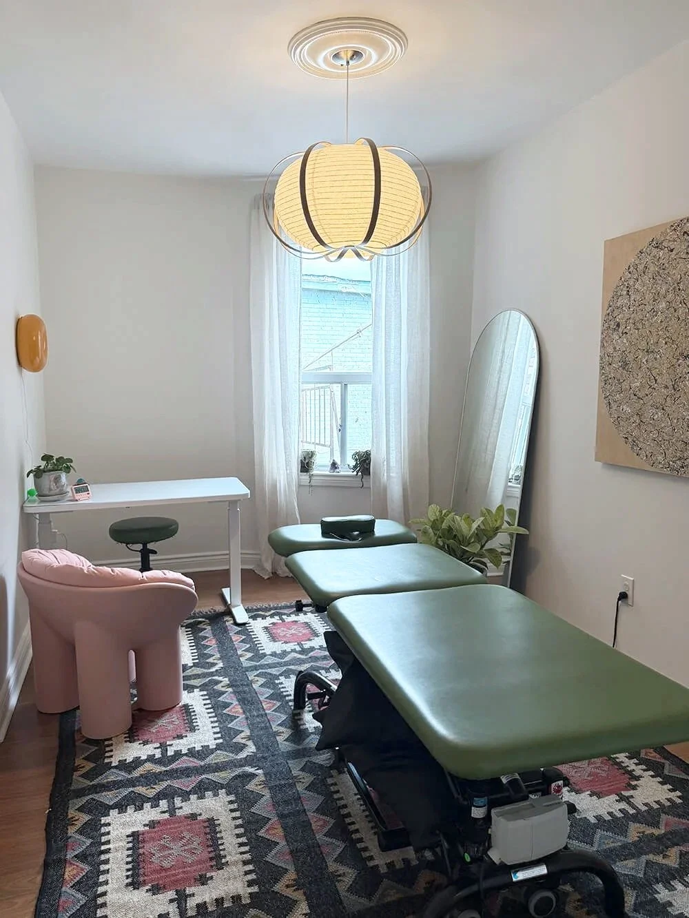A room with a massage table with green covering, a pink animal-shaped armchair, a small white desk with a green stool, a large mirror, a window with sheer white curtains, a wall art piece, and a patterned rug.