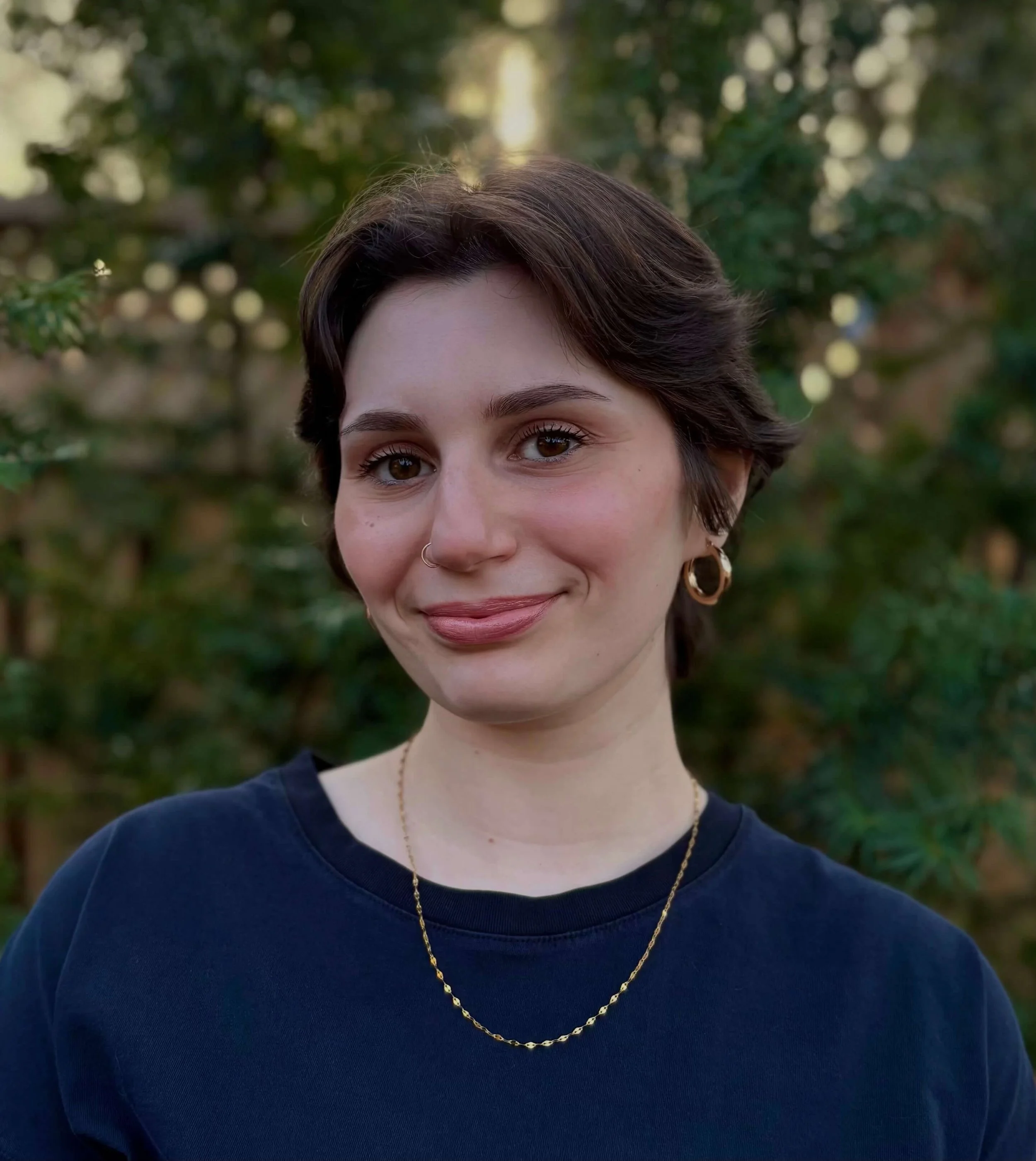 Close-up photo of a young woman with short brown hair, brown eyes, wearing a navy blue top, gold hoop earrings, a gold chain necklace, and a small nose ring, outdoors with blurred green foliage in the background.