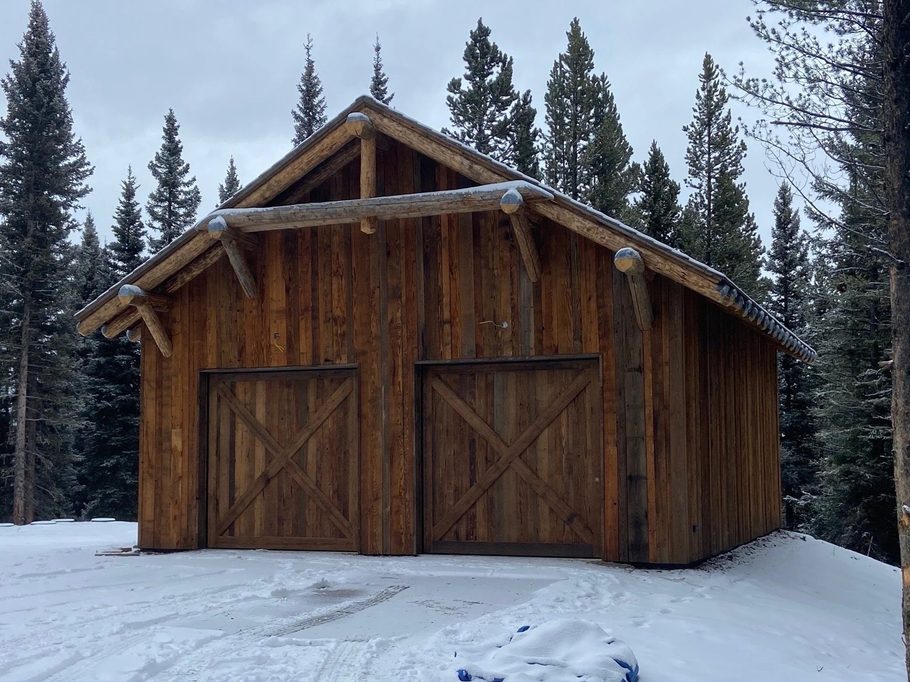 A wooden barn in a snowy forest with tall pine trees in the background.