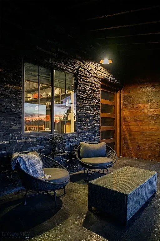 Outdoor patio area with two modern black chairs with pillows, a table, a window reflecting the sunset, and a partially open wooden door, illuminated by warm lighting.