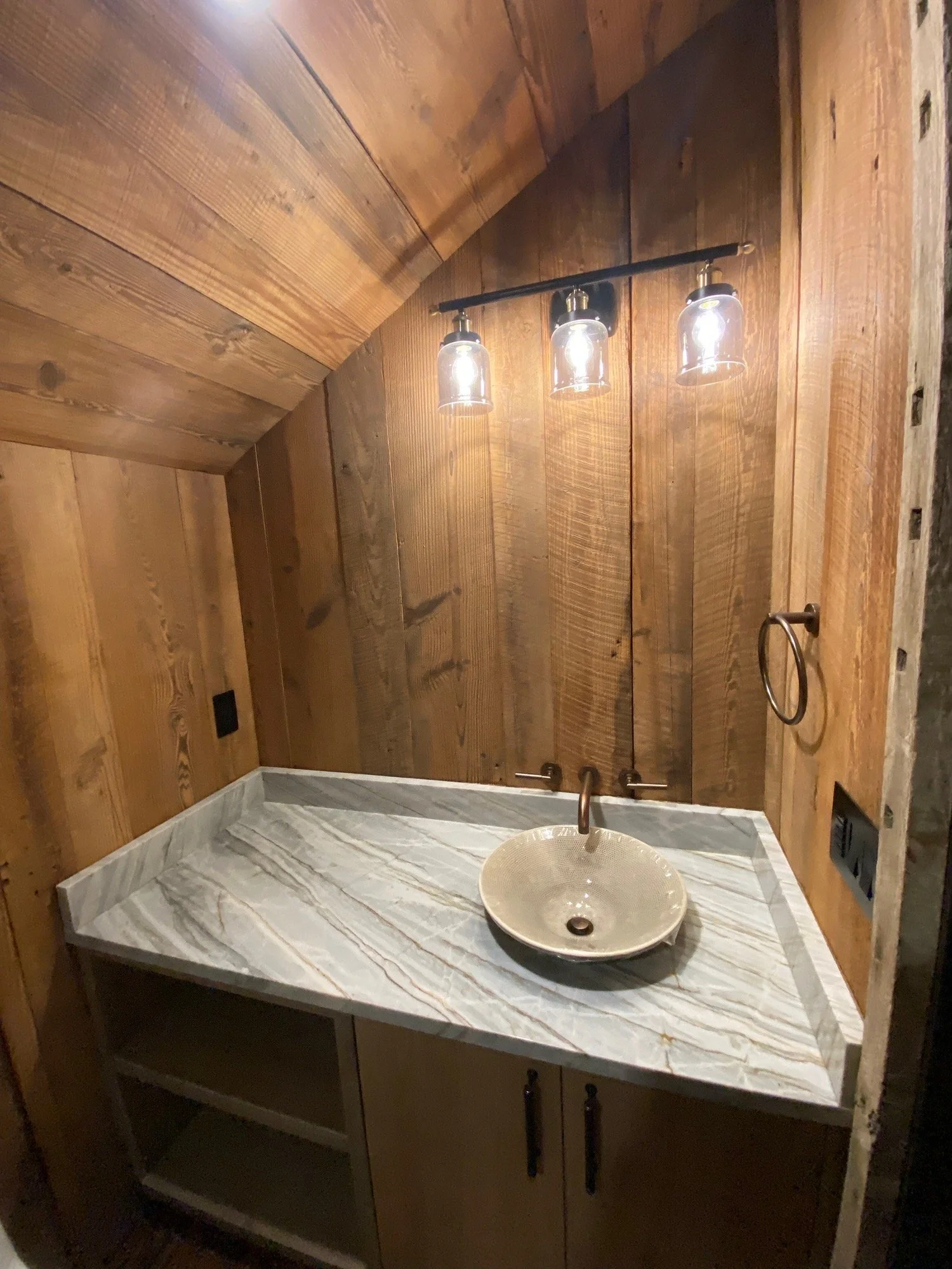 Bathroom with wood-paneled walls and a marble countertop with a vessel sink, illuminated by three glass-encased light bulbs above.