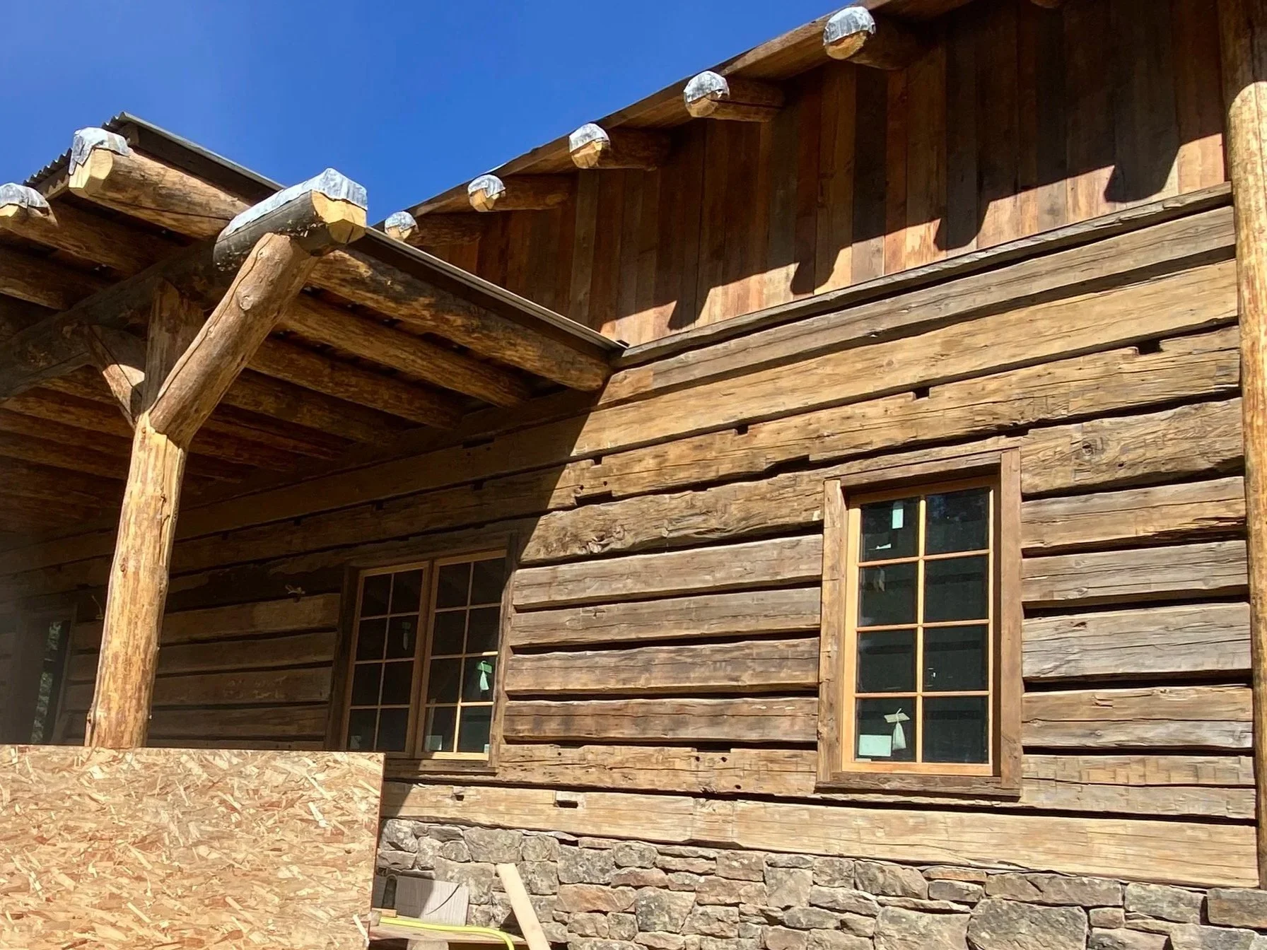 Close-up of a rustic wooden house with two windows, stone foundation, and weathered wood siding, under a clear blue sky.