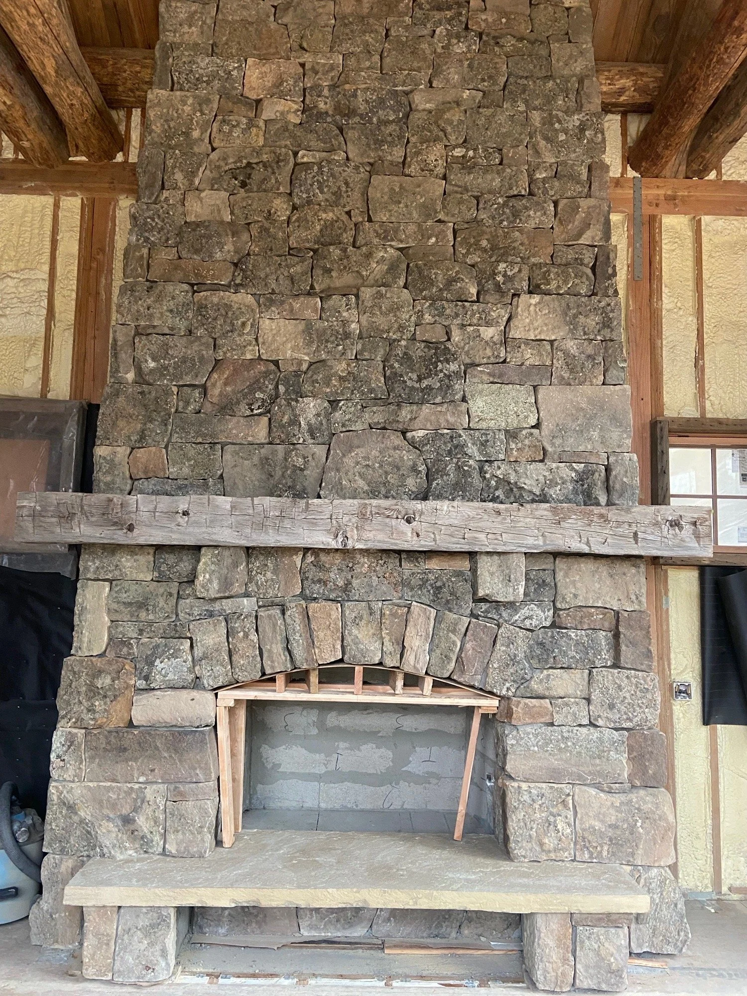 A large stone fireplace under construction with a raw stone chimney, a wooden hearth mantel, and a stone hearth base inside a house under renovation.