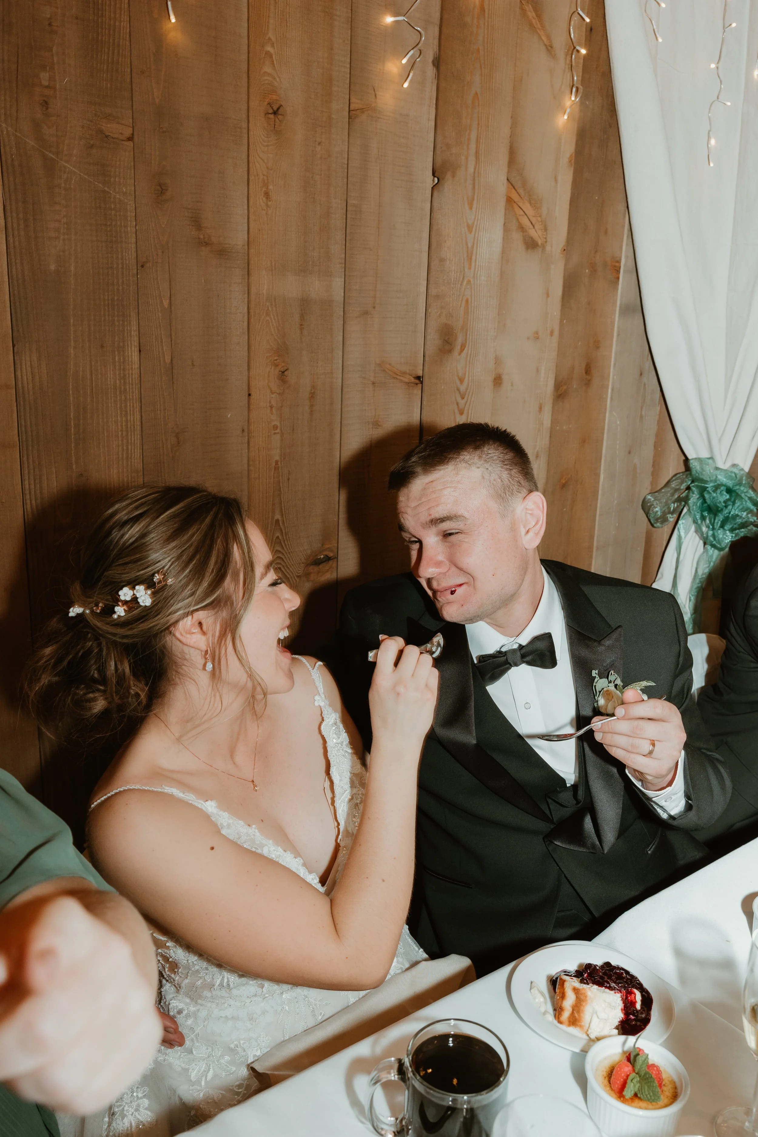Bride and groom sharing a joyful moment during their wedding reception. The bride has a floral hair accessory and is laughing as the groom feeds her. They are dressed in wedding attire and sitting at a table with dessert and drinks.