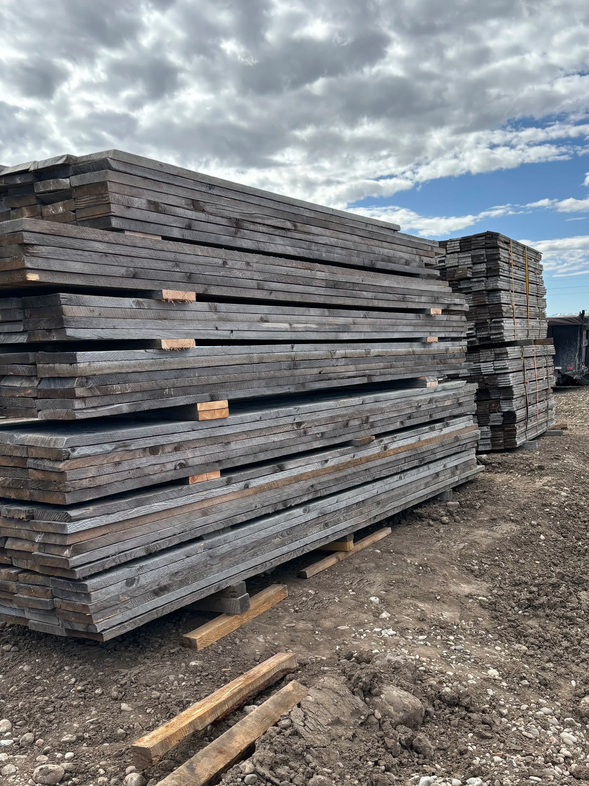 Stacks of wooden planks on a dirt ground with a cloudy sky in the background.