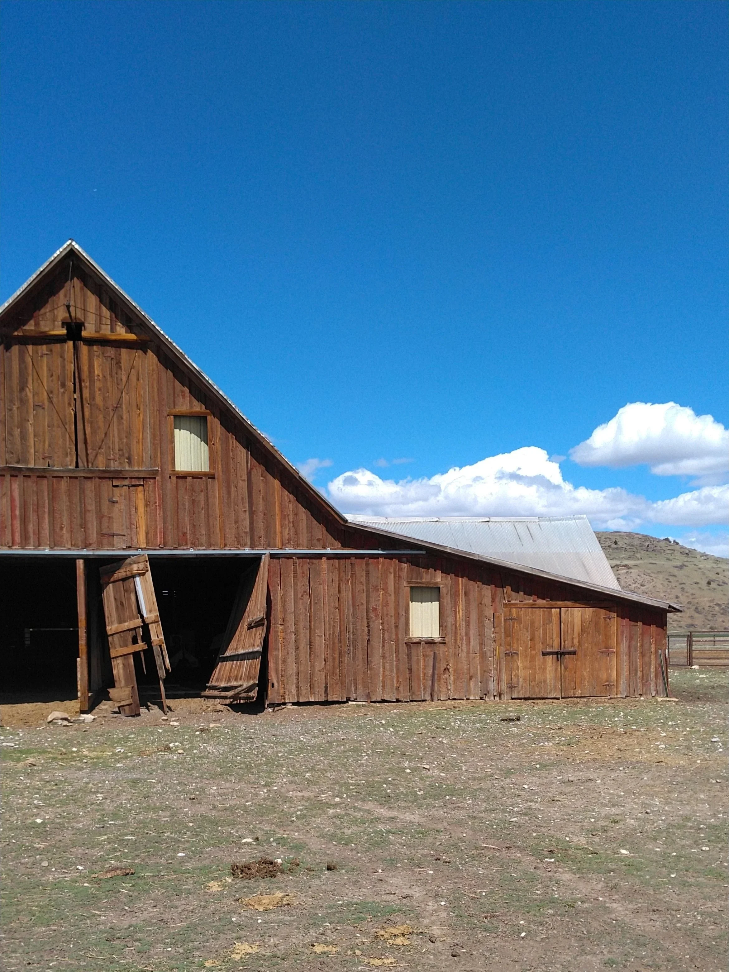 Old wooden barn with a sloped metal roof and open double doors, situated on a flat dirt field under a bright blue sky with clouds and distant hills.