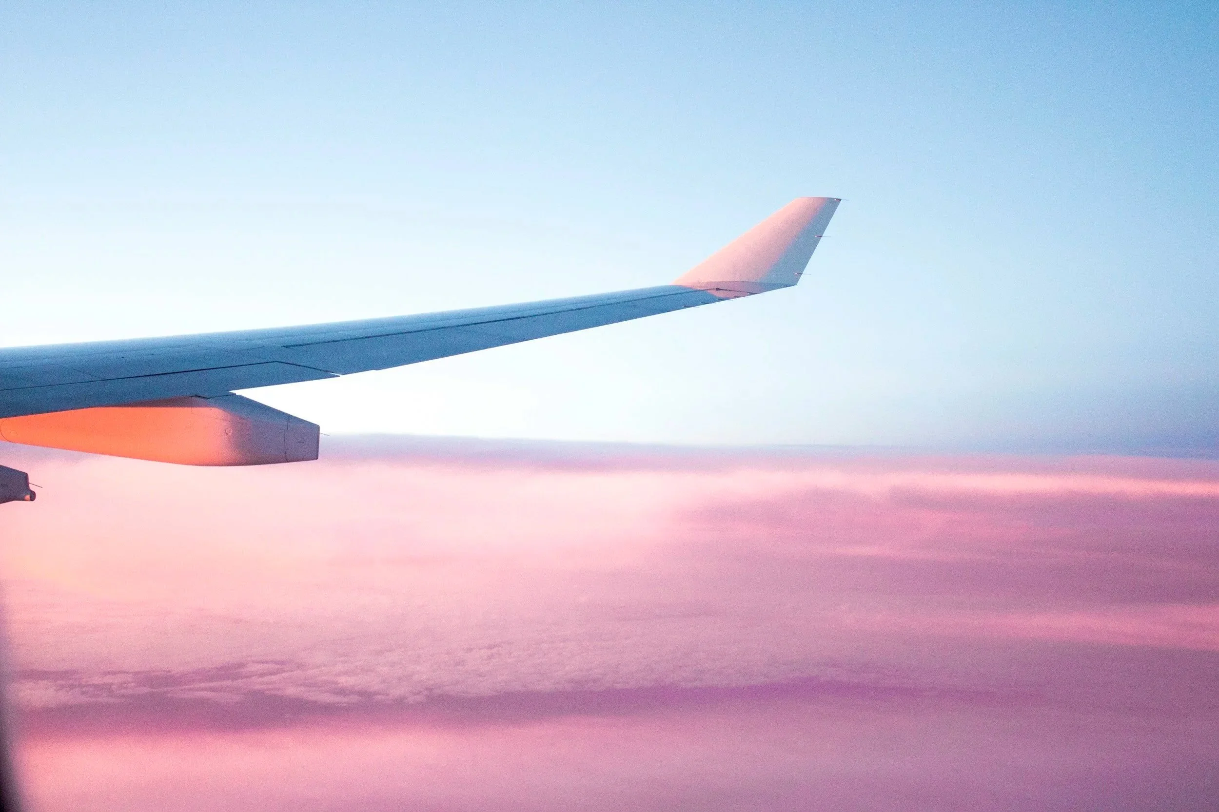 View of an airplane wing in the sky during sunset or sunrise, with a gradient of pink, purple, and blue colors and clouds below.