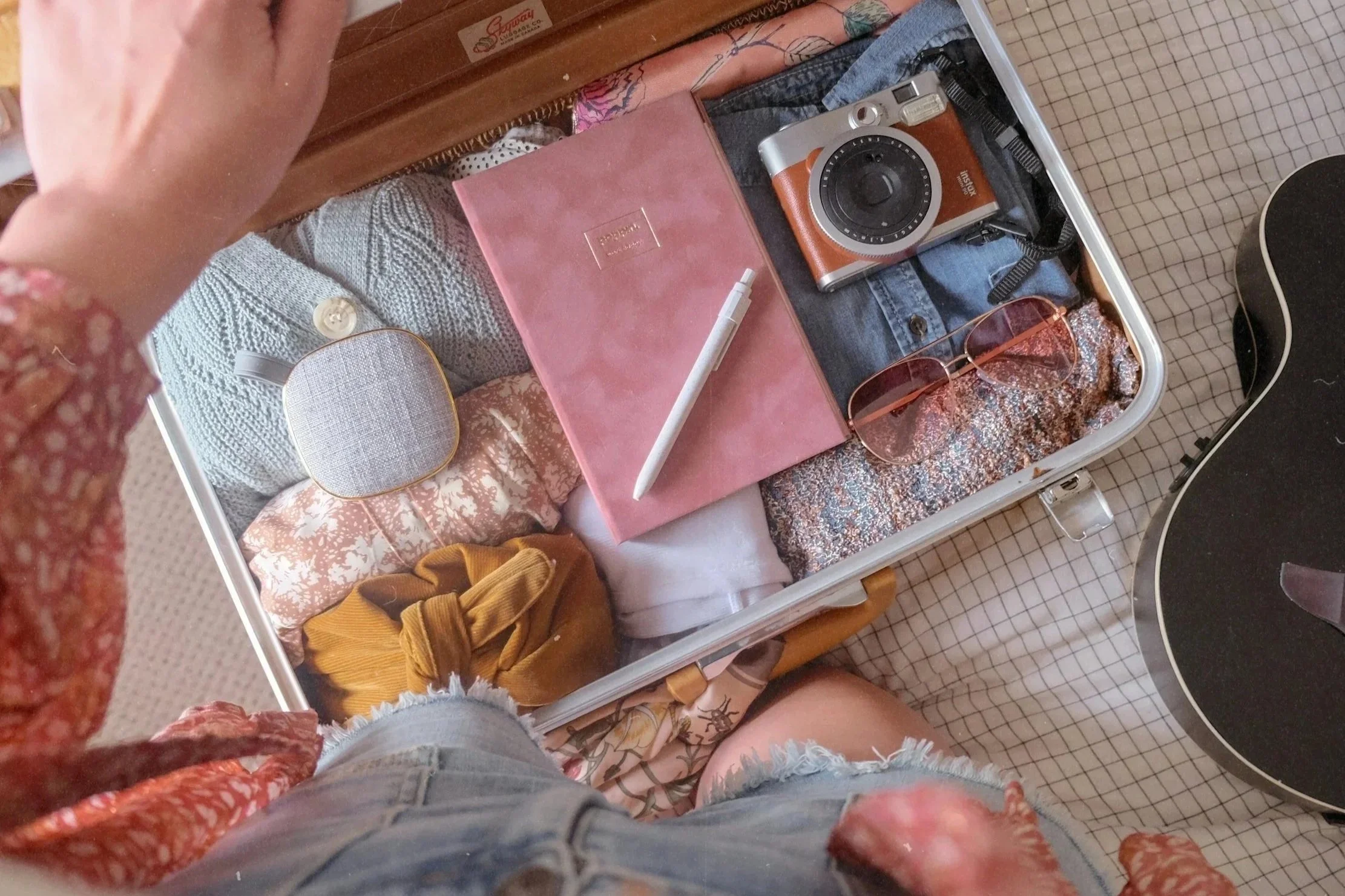 A suitcase open on the floor with various personal items including a camera, pink notebook, sunglasses, a small speaker, and clothing. The photo is taken from above, showing a person's legs and part of their hand at the top of the image.