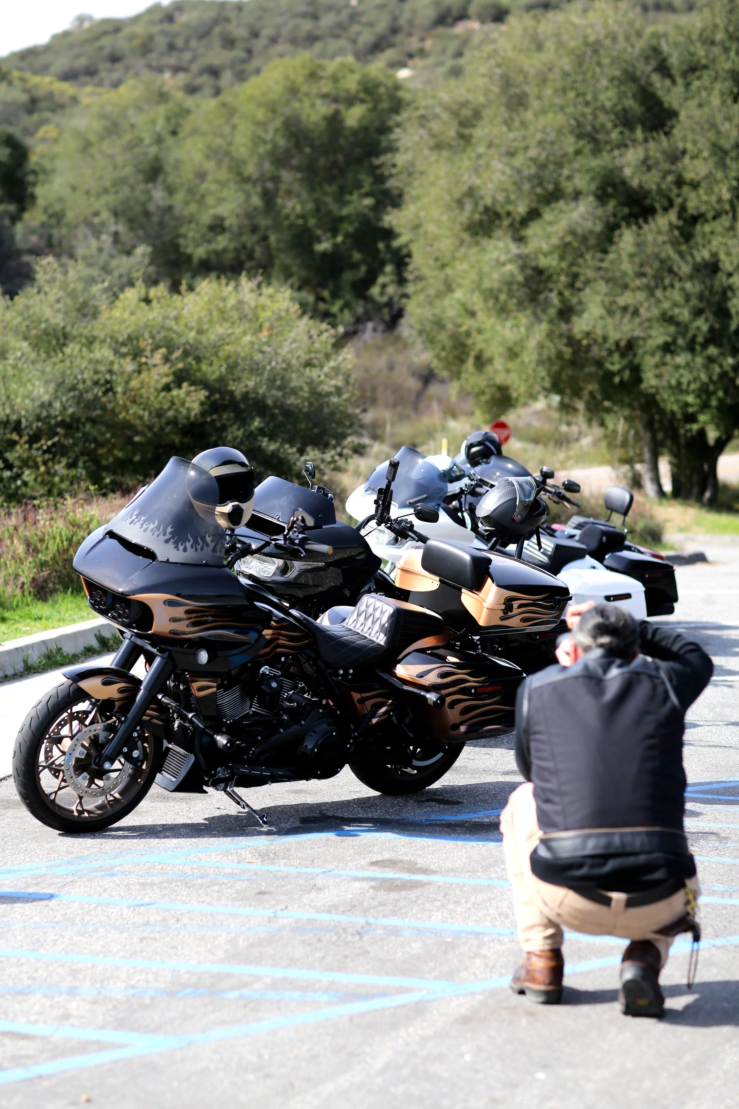 A person in a black jacket crouching in a parking lot taking a photo of a black motorcycle with flame designs. Several other motorcycles with helmets are parked nearby, with greenery and trees in the background.