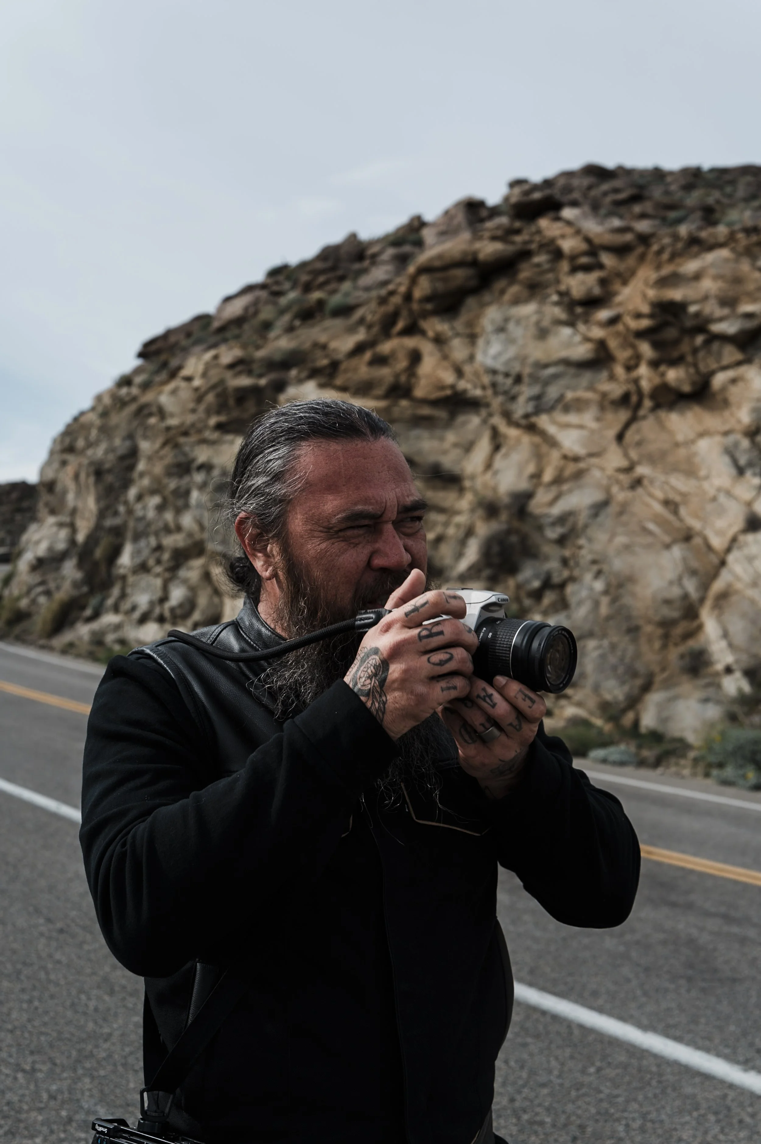 A man with tattoos on his fingers and a beard, dressed in black, is taking a photo with a camera on the roadside with rocky terrain in the background.