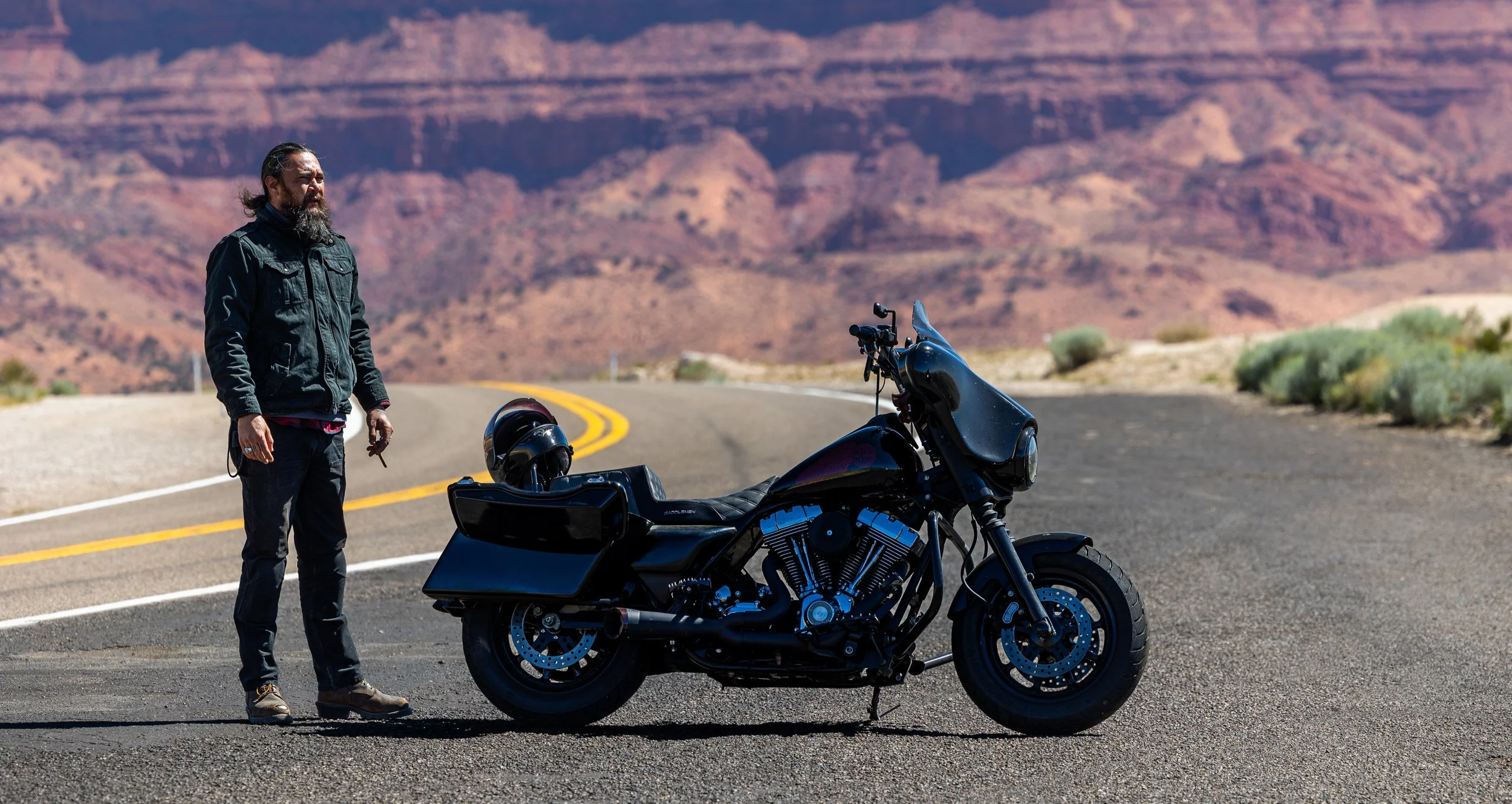 A man with long hair and beard, wearing a black jacket, standing next to a black motorcycle on a winding desert road with colorful rocky hills in the background. A helmet is placed on the motorcycle's rear storage compartment.