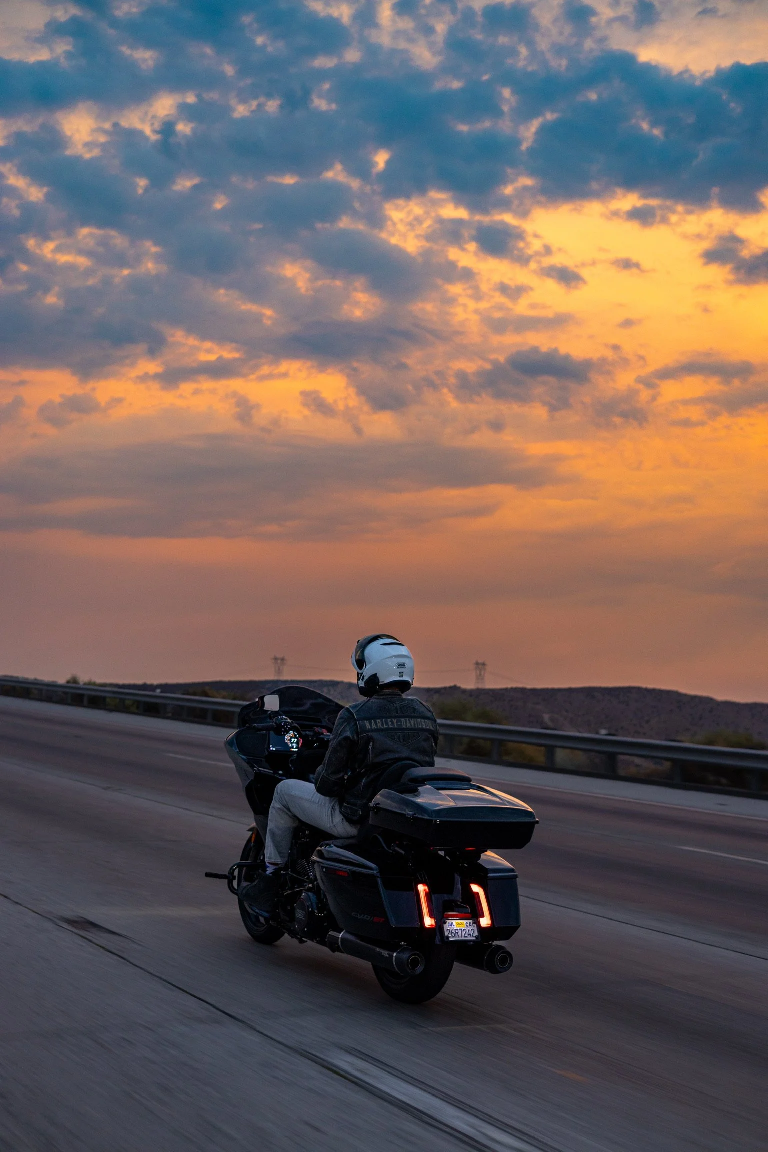 A motorcyclist wearing a helmet and Harley-Davidson jacket riding a black motorcycle on a highway at sunset.
