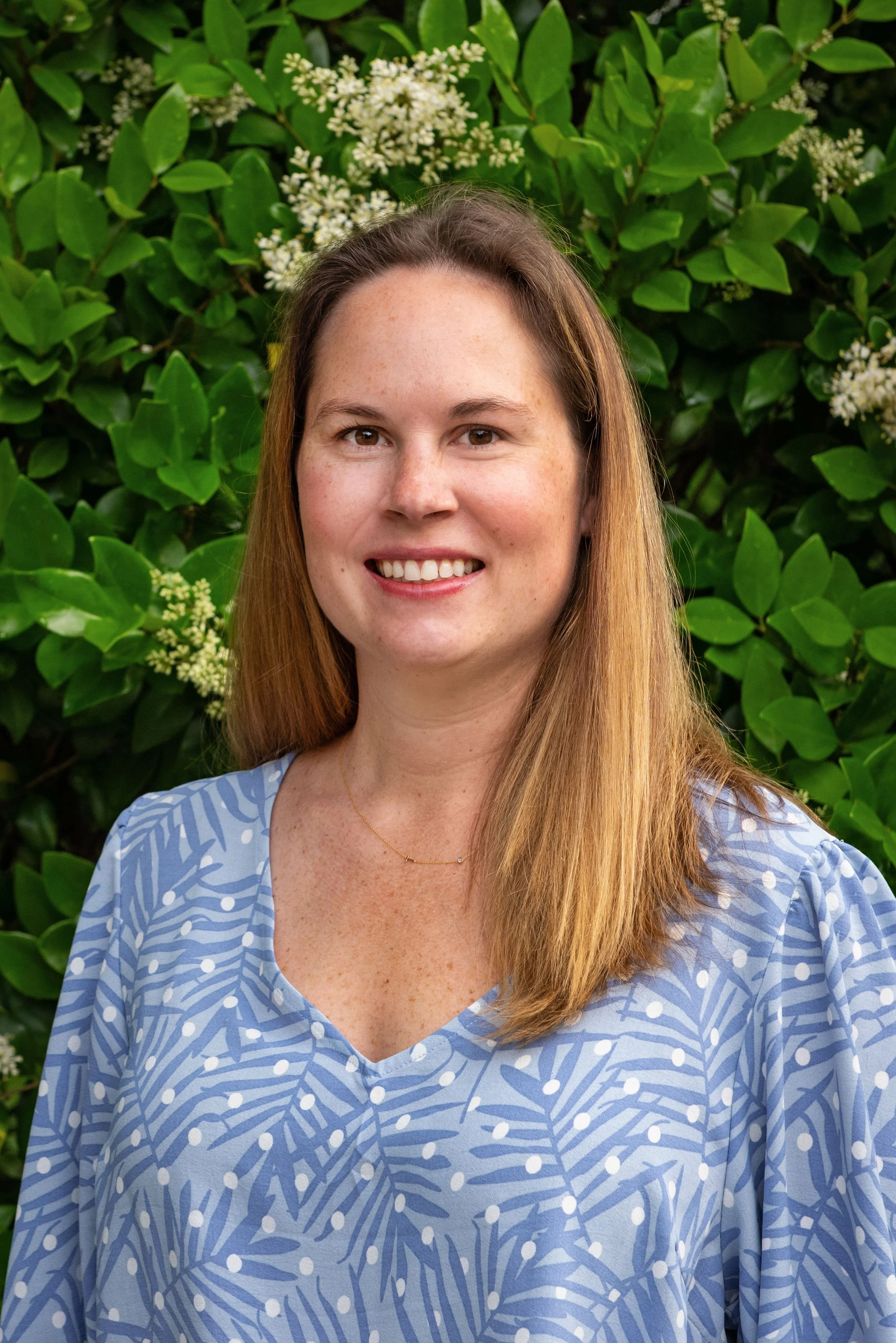 A woman with long, light brown hair smiling, wearing a blue patterned dress, standing outdoors in front of green shrubbery with white flowers.