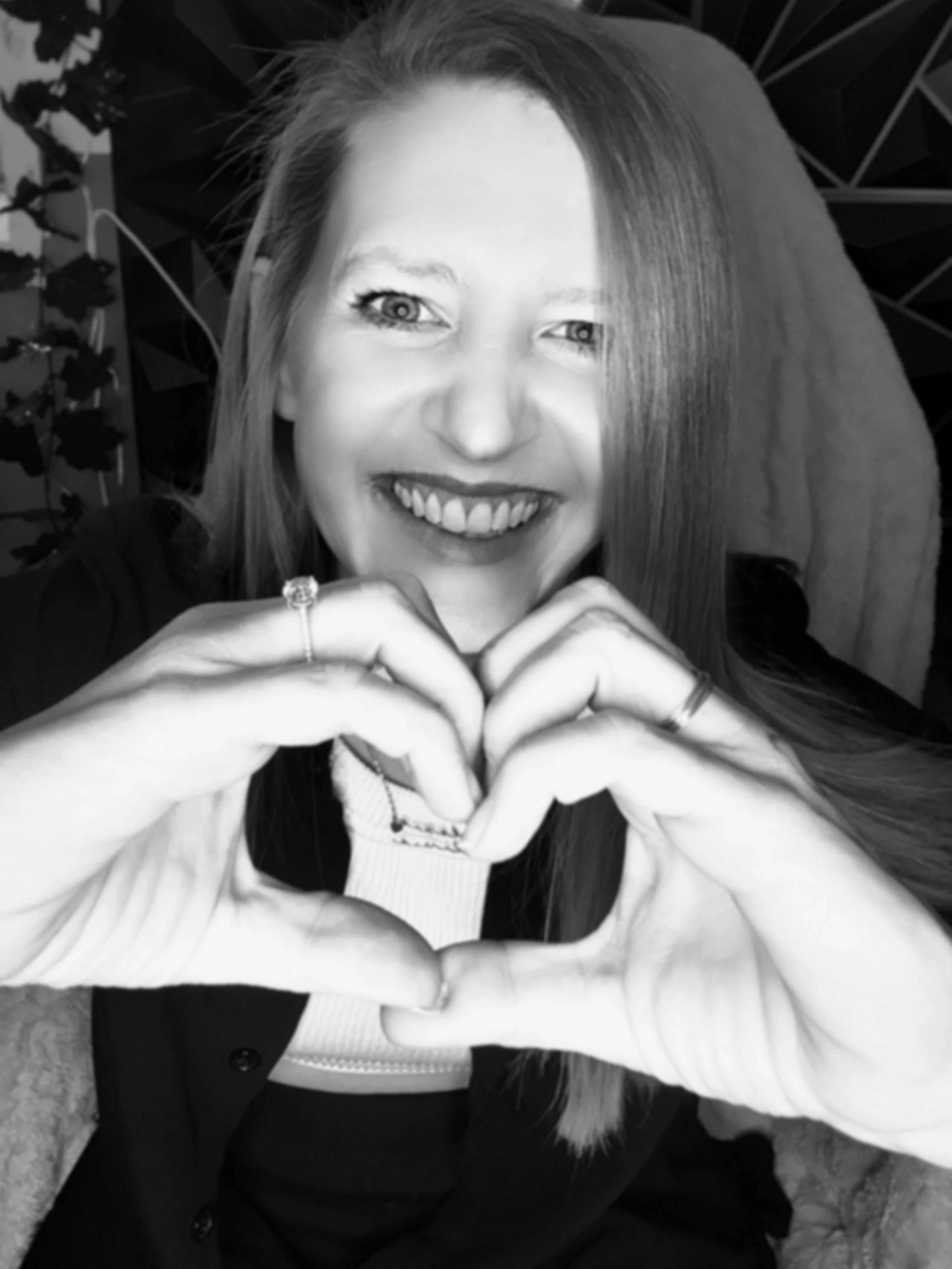 A woman with long hair smiling and forming a heart shape with her hands, with a neon sign in the background that reads 'Willow Refuge'.