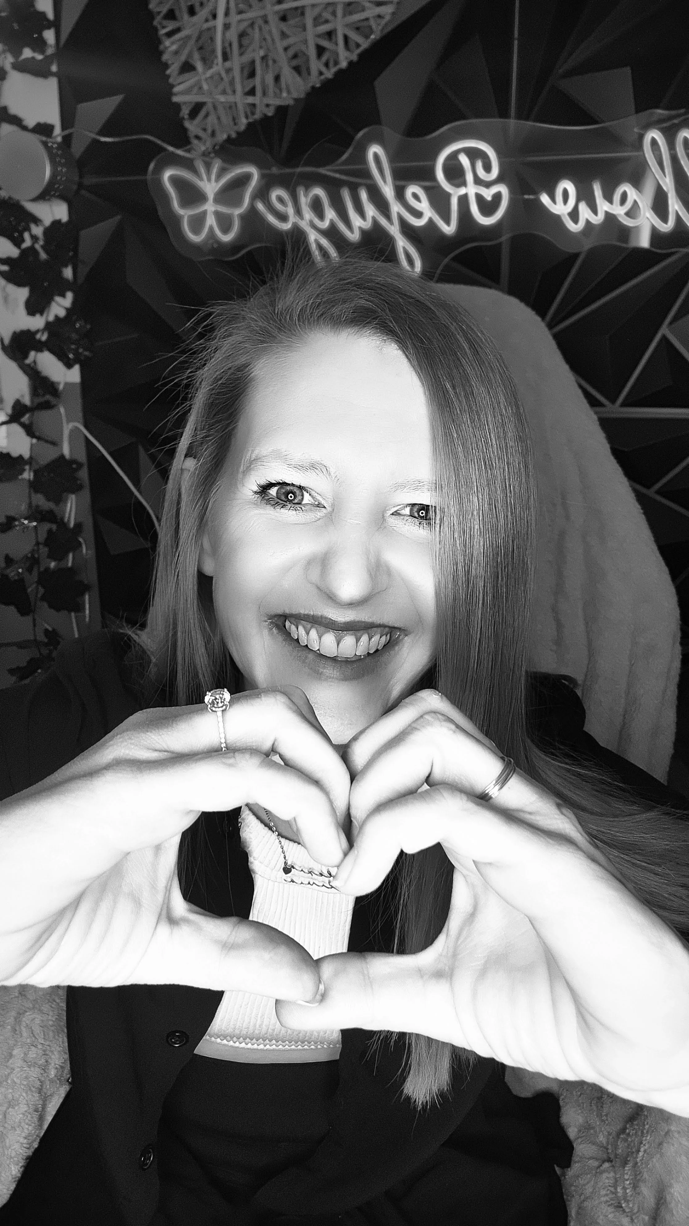 A woman with long hair smiling and forming a heart shape with her hands, with a neon sign in the background that reads 'Willow Refuge'.