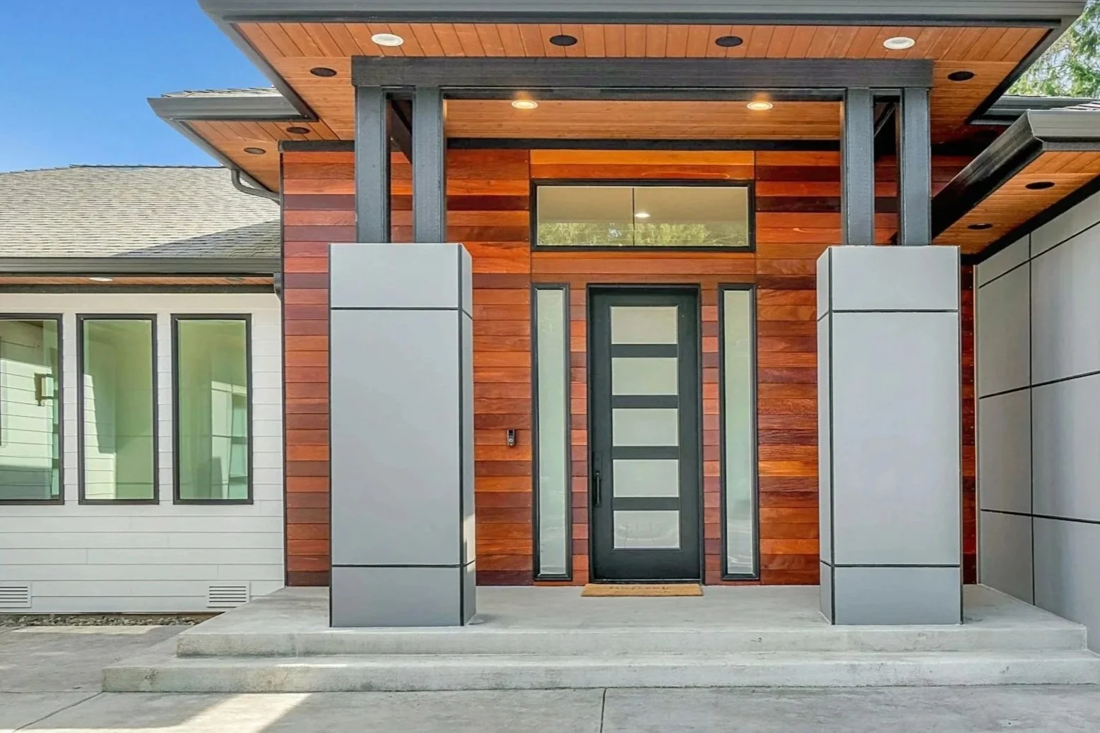 Modern house entrance with a black door, wooden exterior, and gray metal accents, steps leading up to the doorway, and a small welcome mat.