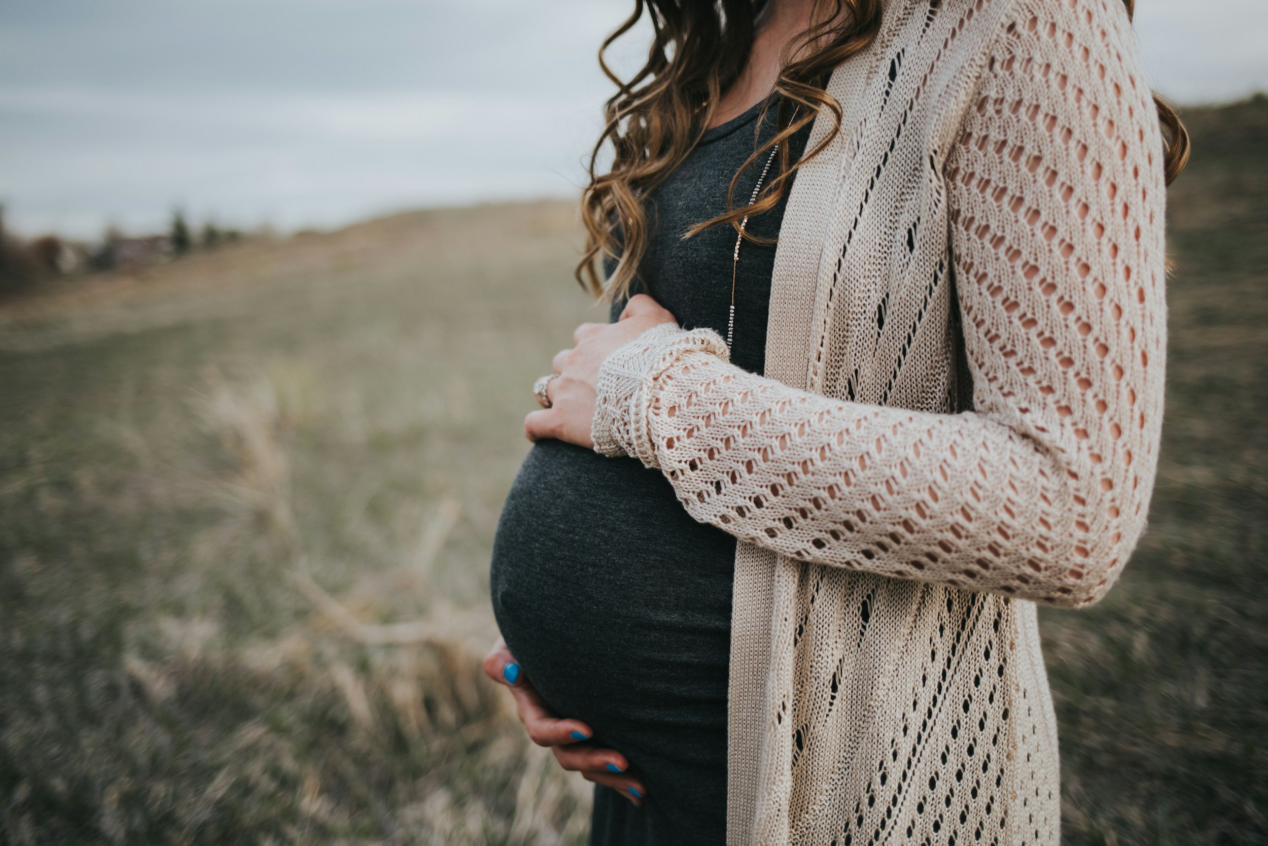 Close-up of pregnant woman outdoors cradling her belly with her hands, wearing a dark shirt and a beige open-knit cardigan.