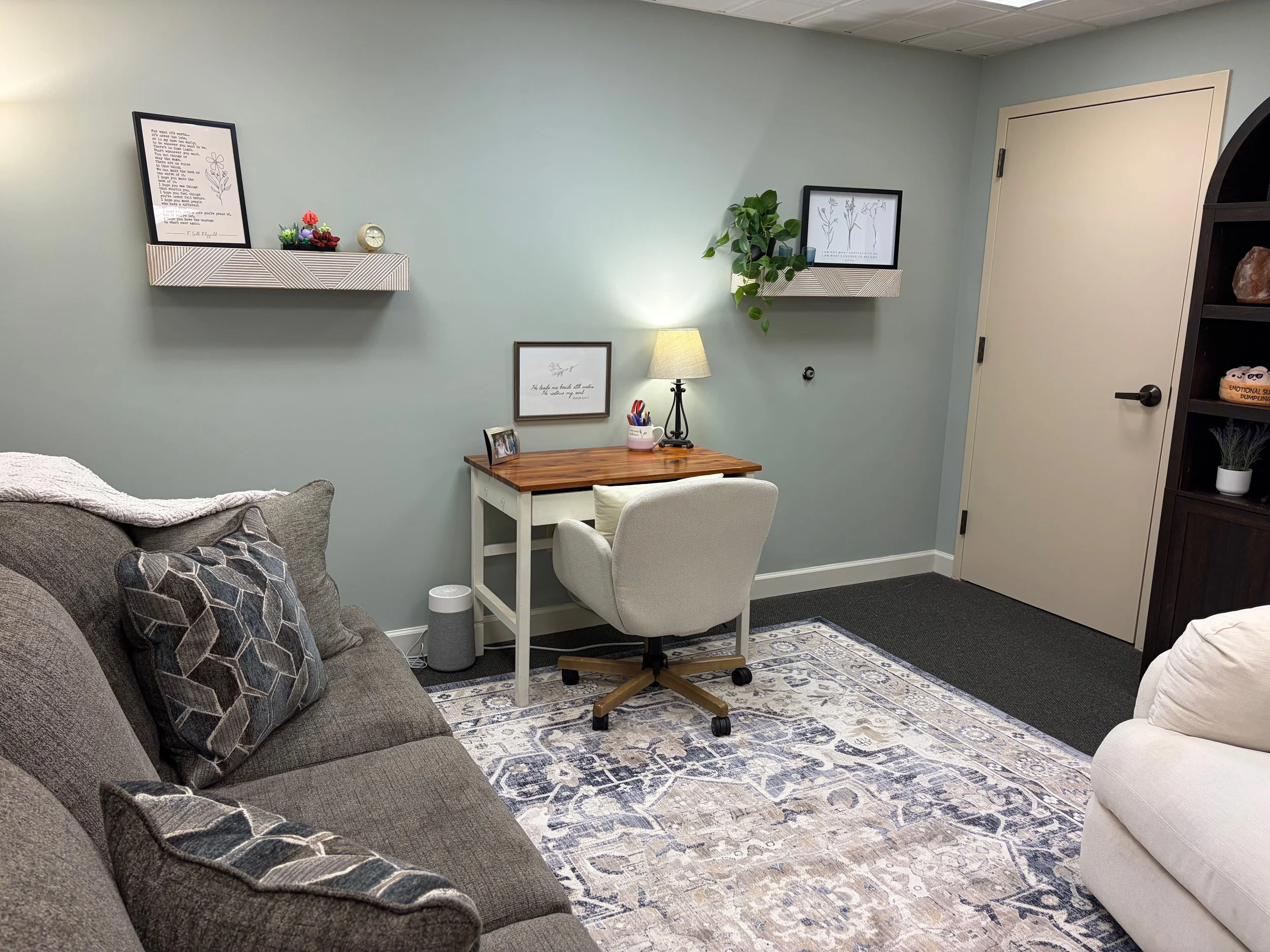 A cozy room with a gray sofa decorated with patterned pillows, a wooden desk with a white office chair, a small lamp, and framed artwork on the walls. There is a patterned area rug on the dark carpeted floor and a bookcase with decorative items. A do