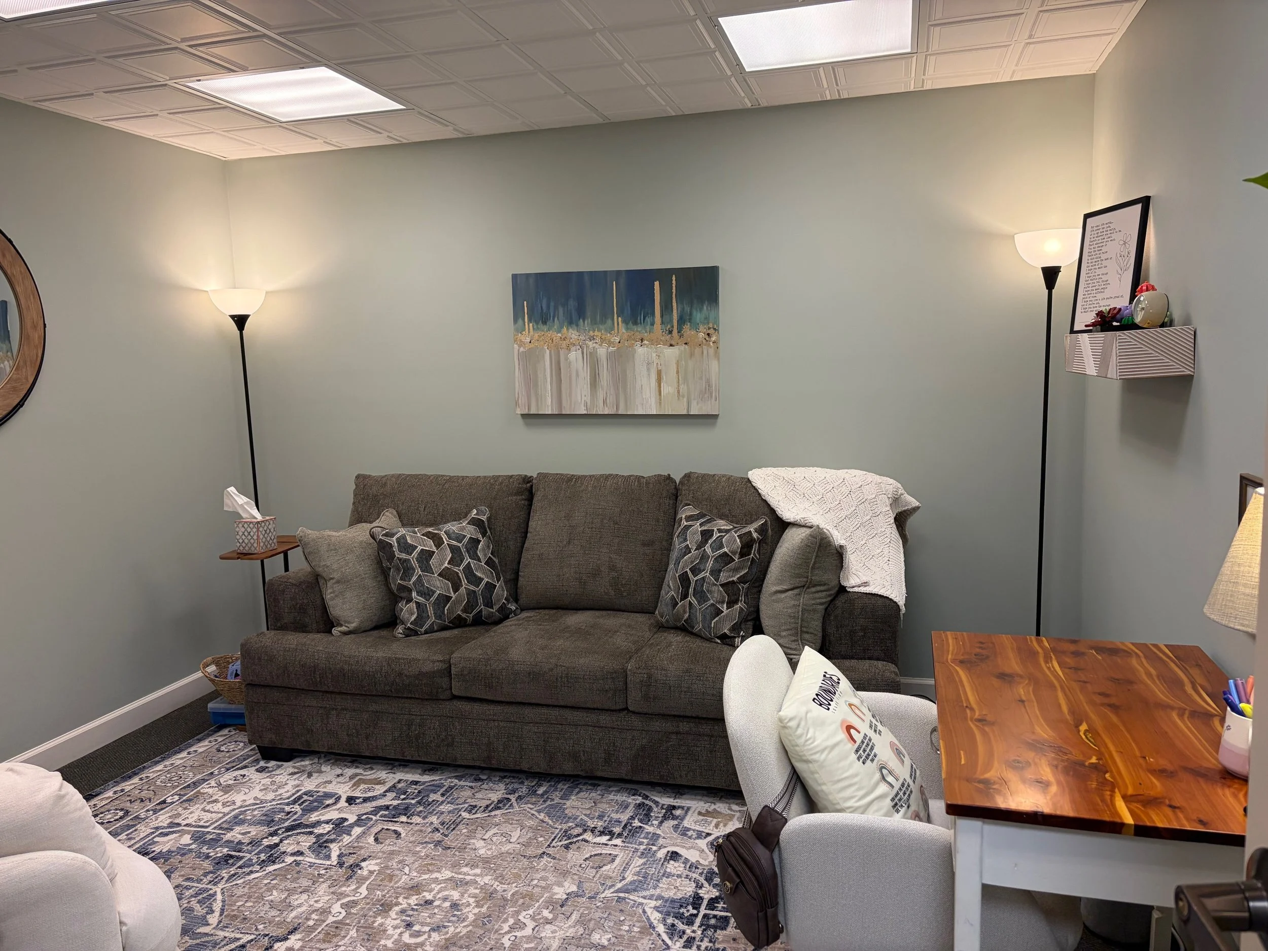 Living room with a gray sofa, patterned pillows, and a white blanket on the right armrest. Wall decorations include abstract art and a framed quote. Two floor lamps provide lighting, and a wooden table with pens and a cushion are in the foreground. A