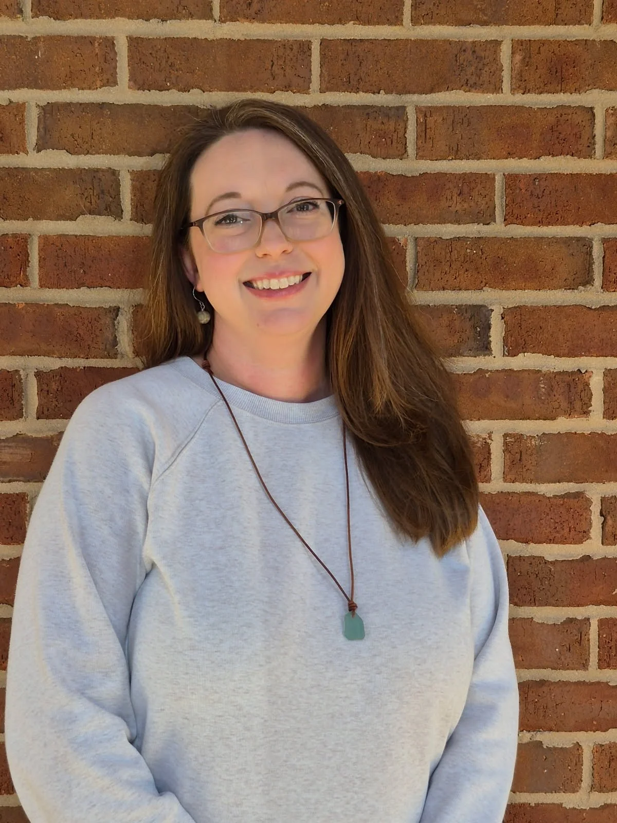 A woman with long brown hair, glasses, and earrings, smiling in front of a brick wall, wearing a light gray sweatshirt and a green pendant necklace.
