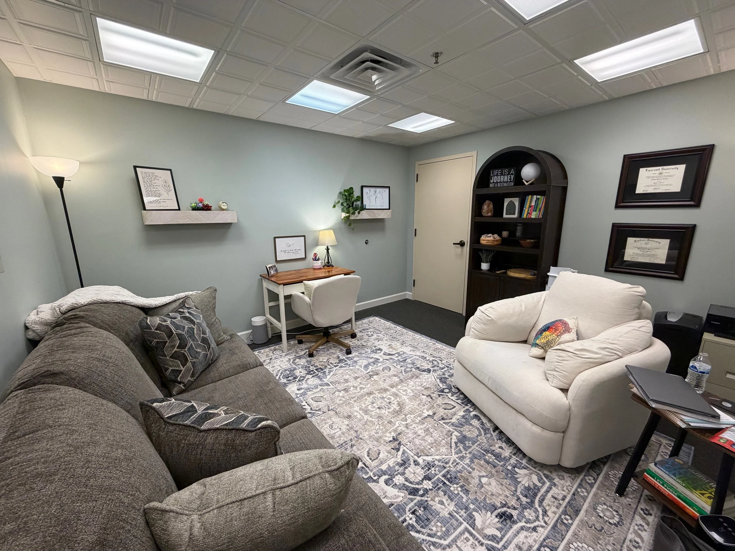 A cozy office or living room with a brown couch with patterned pillows, a cream armchair, a wooden desk with a chair, a dark bookshelf with books and decorative items, framed certificates and art on the walls, a floor lamp, and a ceiling with recesse