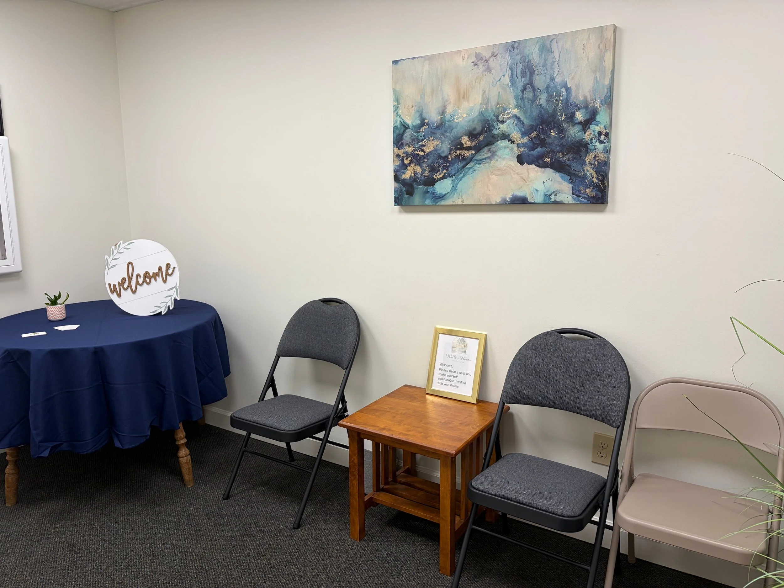 A wall with a modern abstract painting in shades of blue, white, and gold. There are five chairs in front of the wall, a small wooden table with a framed sign and a potted plant, and a table draped with a blue cloth. There is a sign on the table that