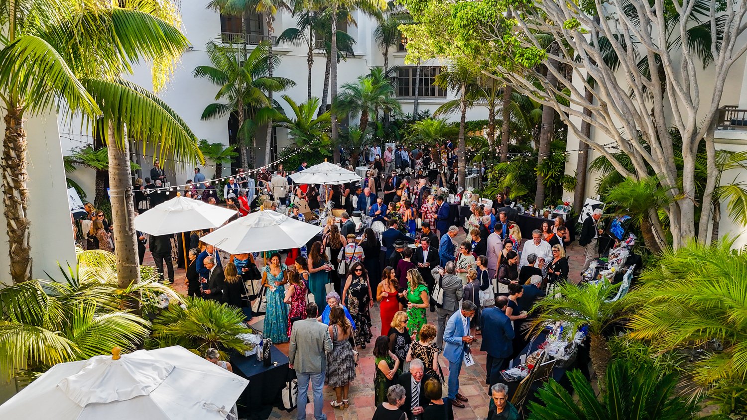 A large outdoor event in a courtyard with many people socializing, surrounded by palm trees and lush greenery, with white umbrellas providing shade.