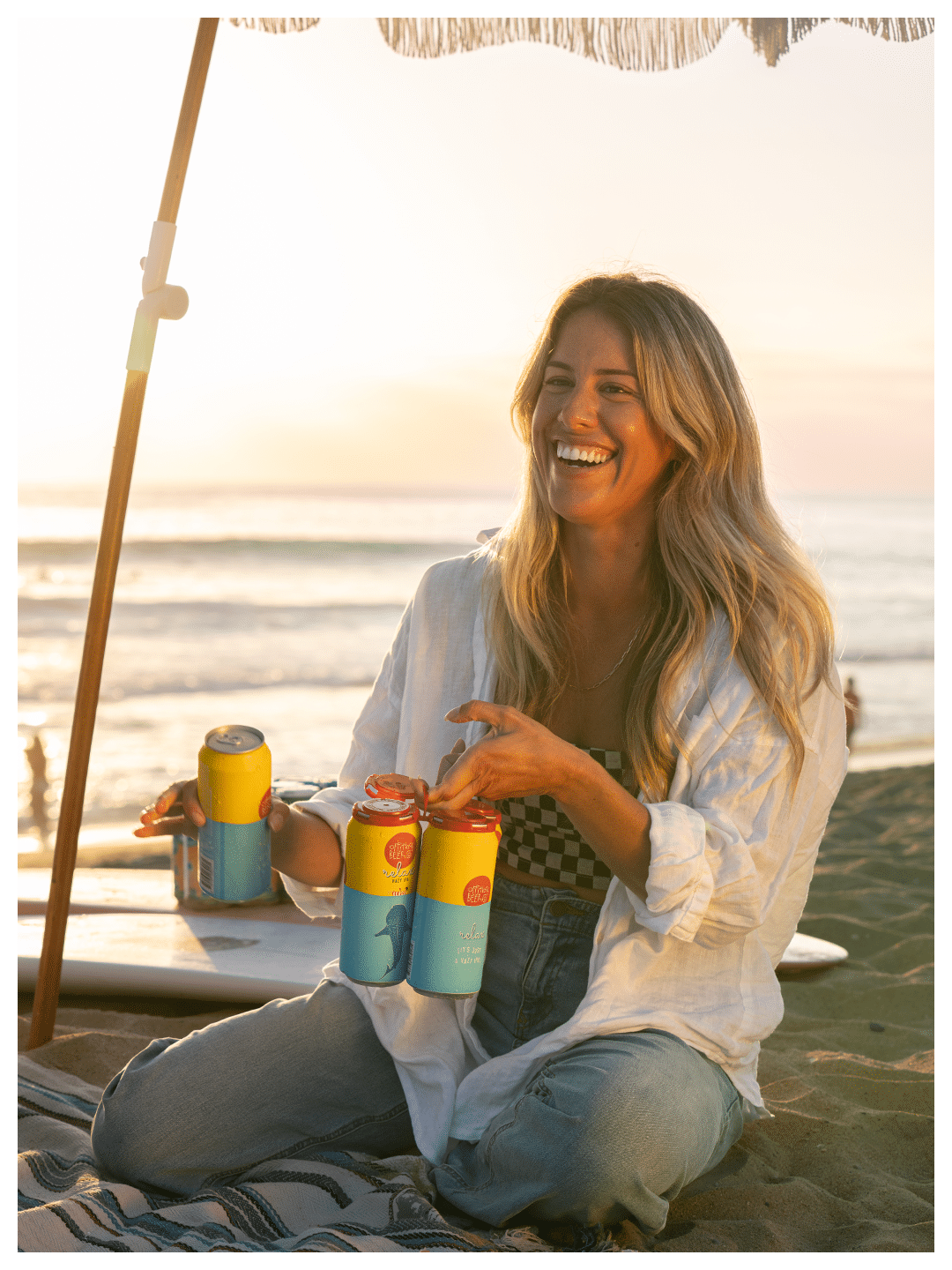 A woman sitting on the beach, smiling, holding four colorful cans, with an umbrella and the ocean in the background at sunset.