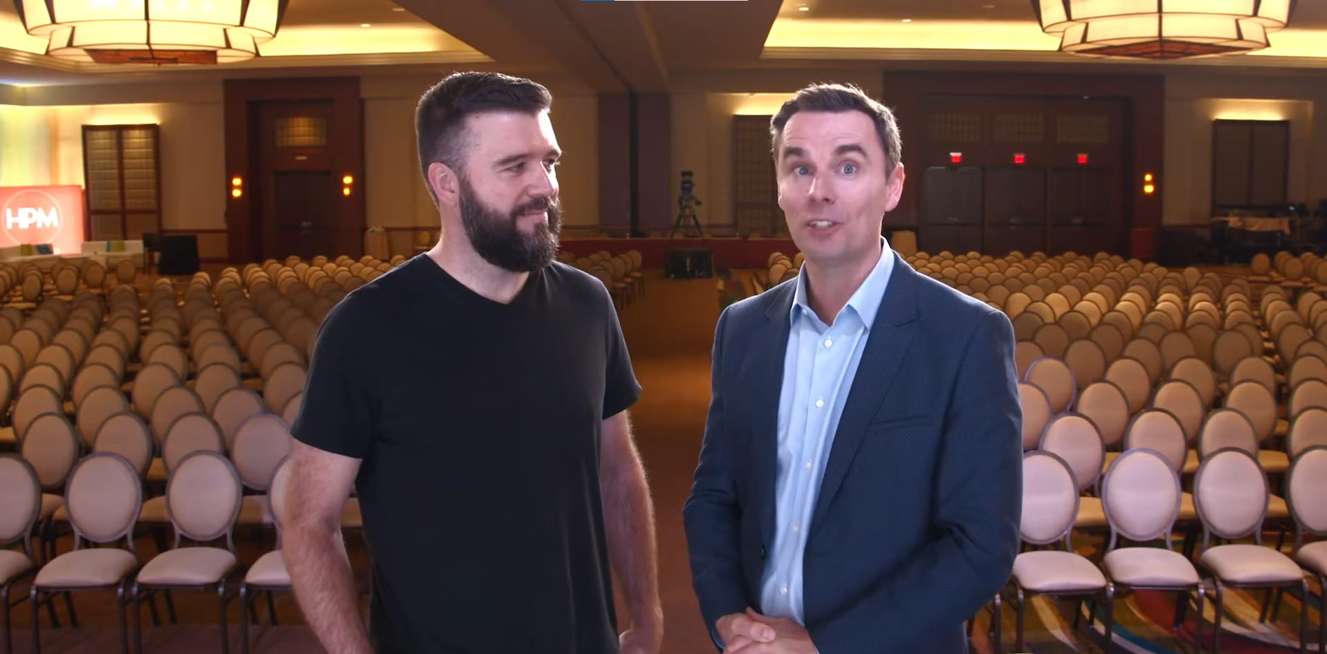 Two men talking in an empty conference hall with rows of beige chairs, soft lighting, and a red screen with white text in the background.