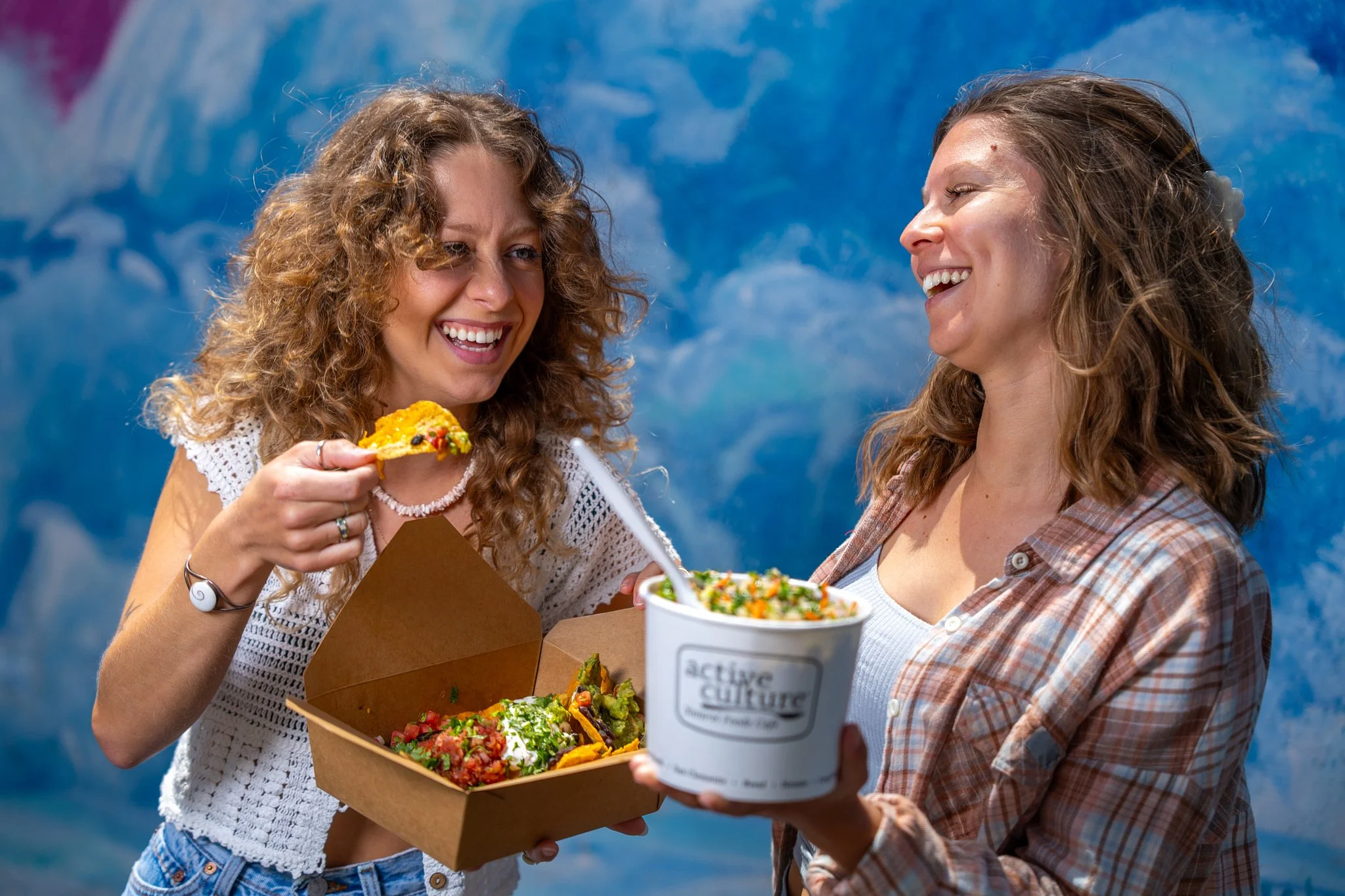 Two women with curly hair enjoying a meal together outdoors, smiling, with bowls and boxes of food in front of them, against a blue background.